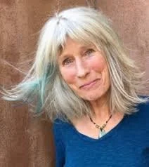 A woman with shoulder-length blonde hair with light blue streaks, wearing a blue top and a necklace, smiling outdoors.