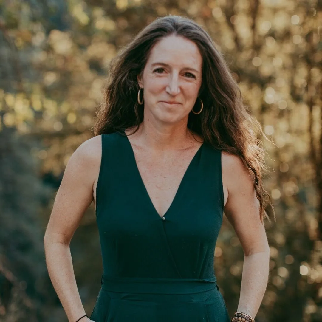 A woman with long, curly brown hair wearing a sleeveless dark green dress and hoop earrings, standing outdoors with a blurred background of trees and autumn foliage.
