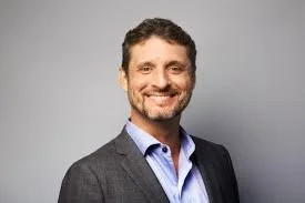 A smiling man with dark hair and a beard, wearing a gray blazer over a light blue shirt, standing against a light gray background.