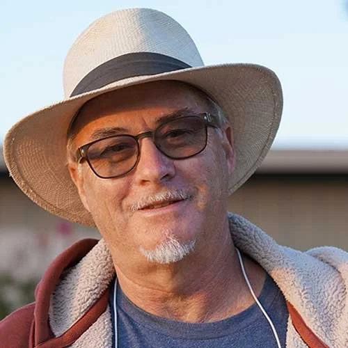 A man wearing a wide-brimmed straw hat, glasses, and a hoodie, smiling outdoors.