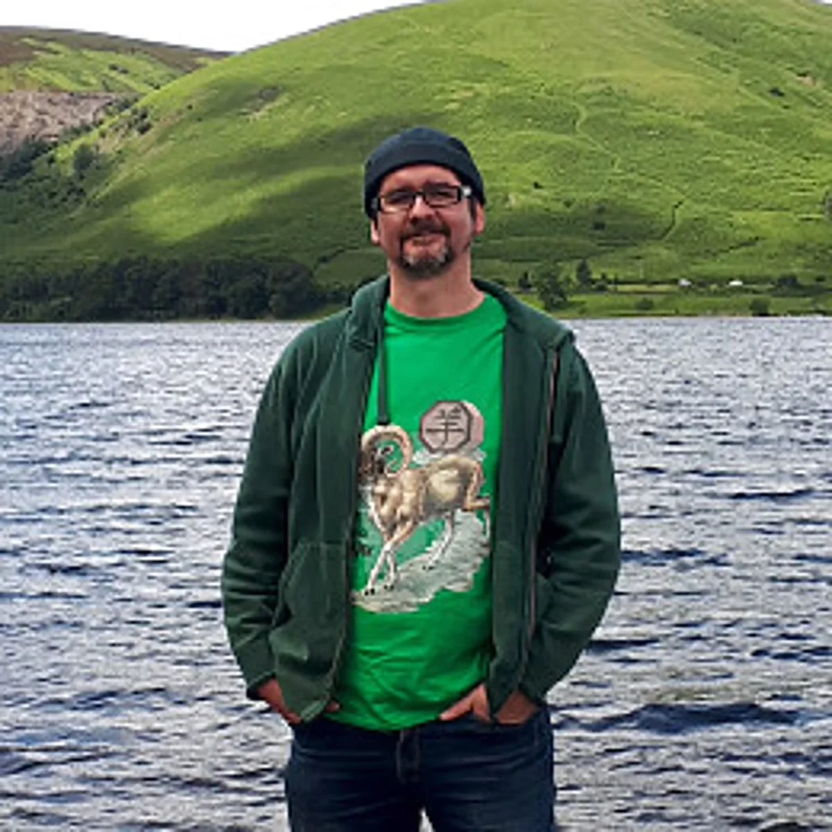 A man standing by a body of water with green hills in the background.