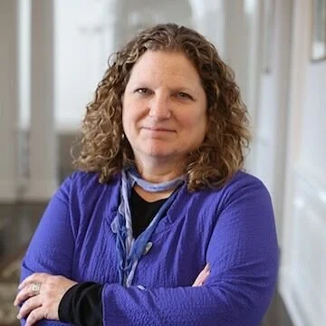 A woman with curly hair wearing a blue sweater and a gray scarf, crossing her arms and standing in a well-lit indoor space.