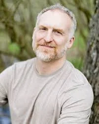 Portrait of a middle-aged man with gray hair and beard, wearing a light gray t-shirt, standing outdoors with trees and greenery in the background.
