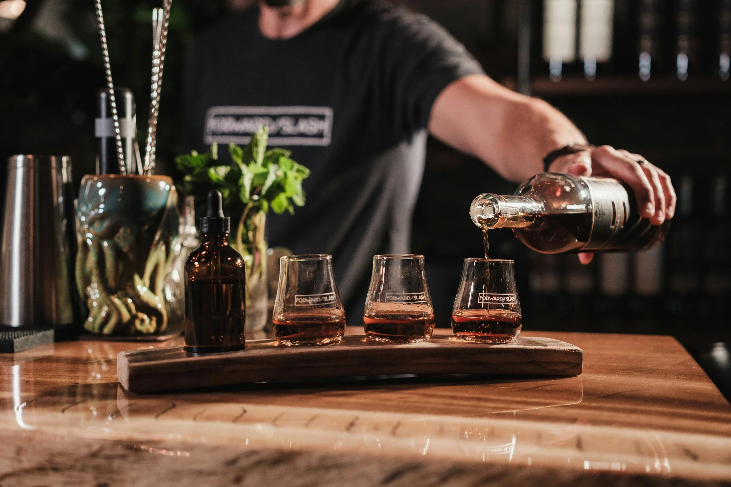 A professional Twyst Cocktail Co. bartender pours a dark spirit into a flight of three tasting glasses on a curved wooden board, surrounded by fresh mint, bitters, and stainless steel bar tools.