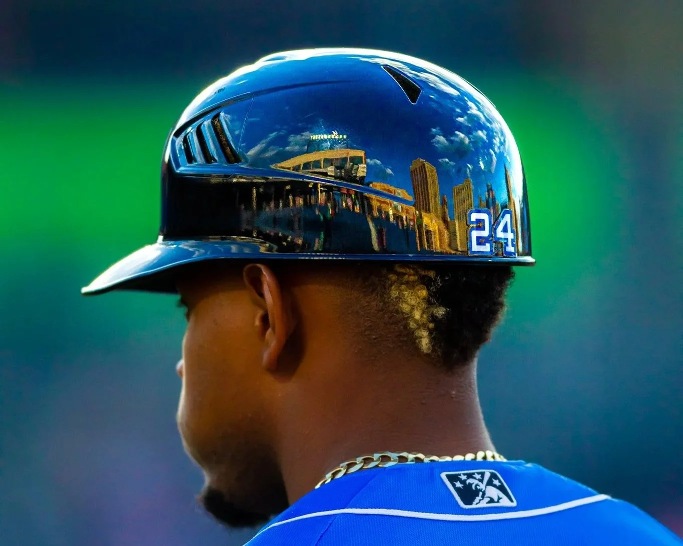 Close-up of a baseball player wearing a shiny blue helmet with a city skyline reflection, in front of a blurred green and blue background.