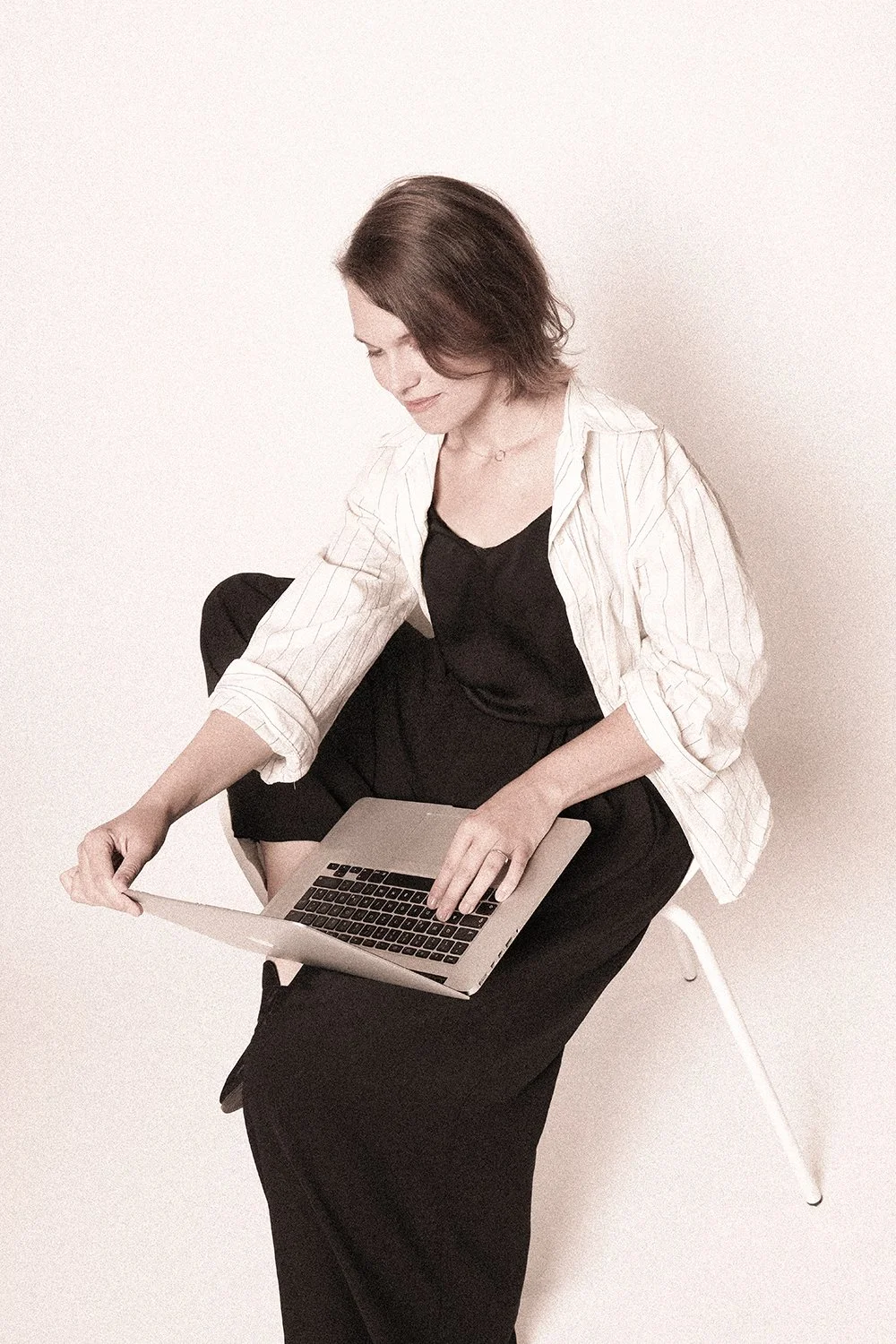 Eine Frau sitzt auf einem weißen Stuhl gegen eine weiße Wand und arbeitet an einem Laptop.