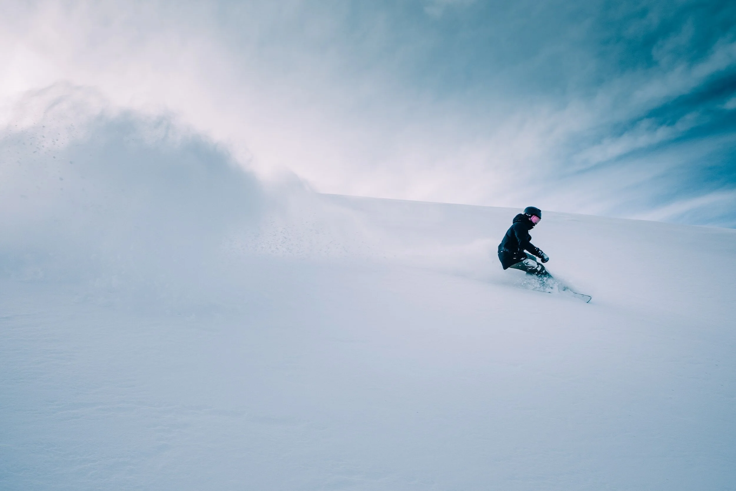 Snødekt fjell, litt skyer på himmelen i bakgrunnen. Person i full fart nedover på snowboard. En vind av snø flyr bak personen.