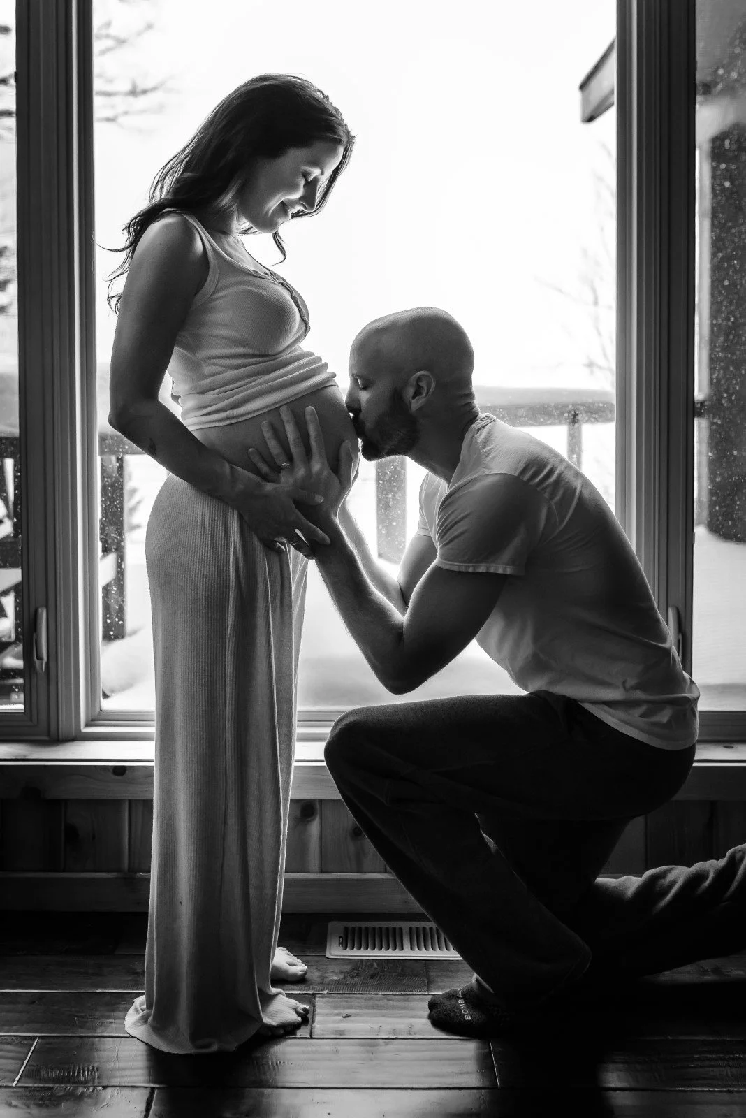 A man kneeling and kissing a pregnant woman's belly inside a house, with a large glass door showing an outdoor view.