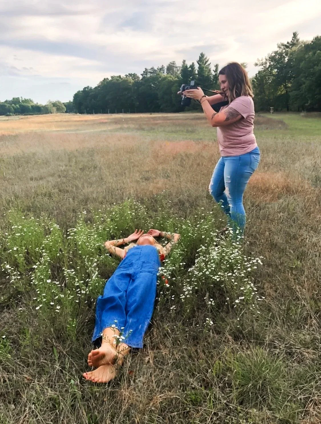 Duluth, Minnesota photographer stands in a field taking a photo of a girl lying on the ground in denim overalls, surrounded by wildflowers.
