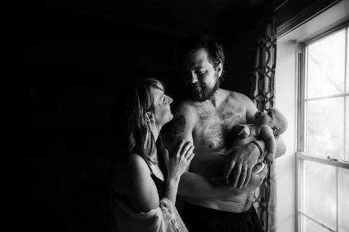 family with newborn stands in front of a nursery window at their home in wausau Wisconsin wall during her maternity session