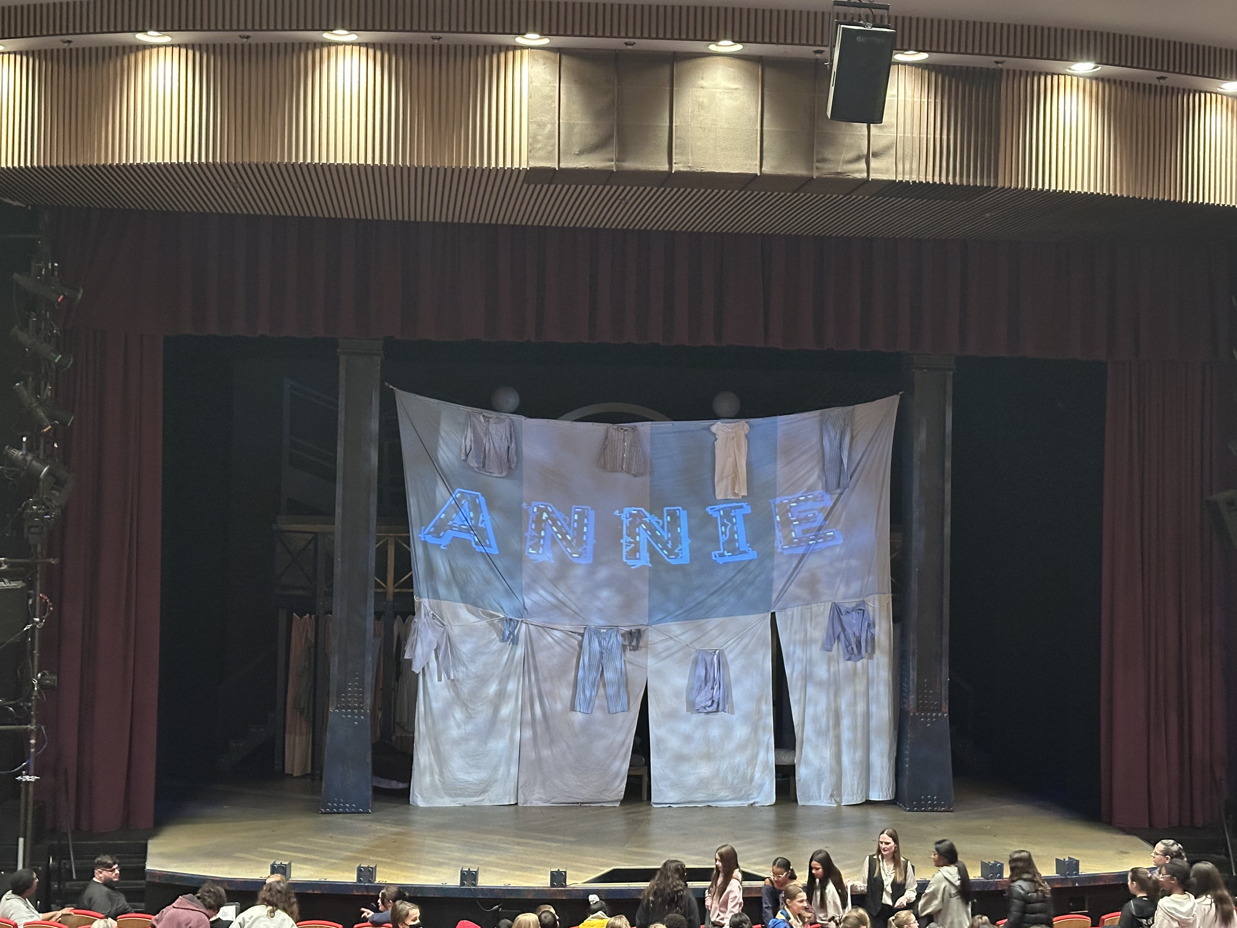 Theater stage with white backdrop displaying 'ANNIE' in blue letters with hanging costumes