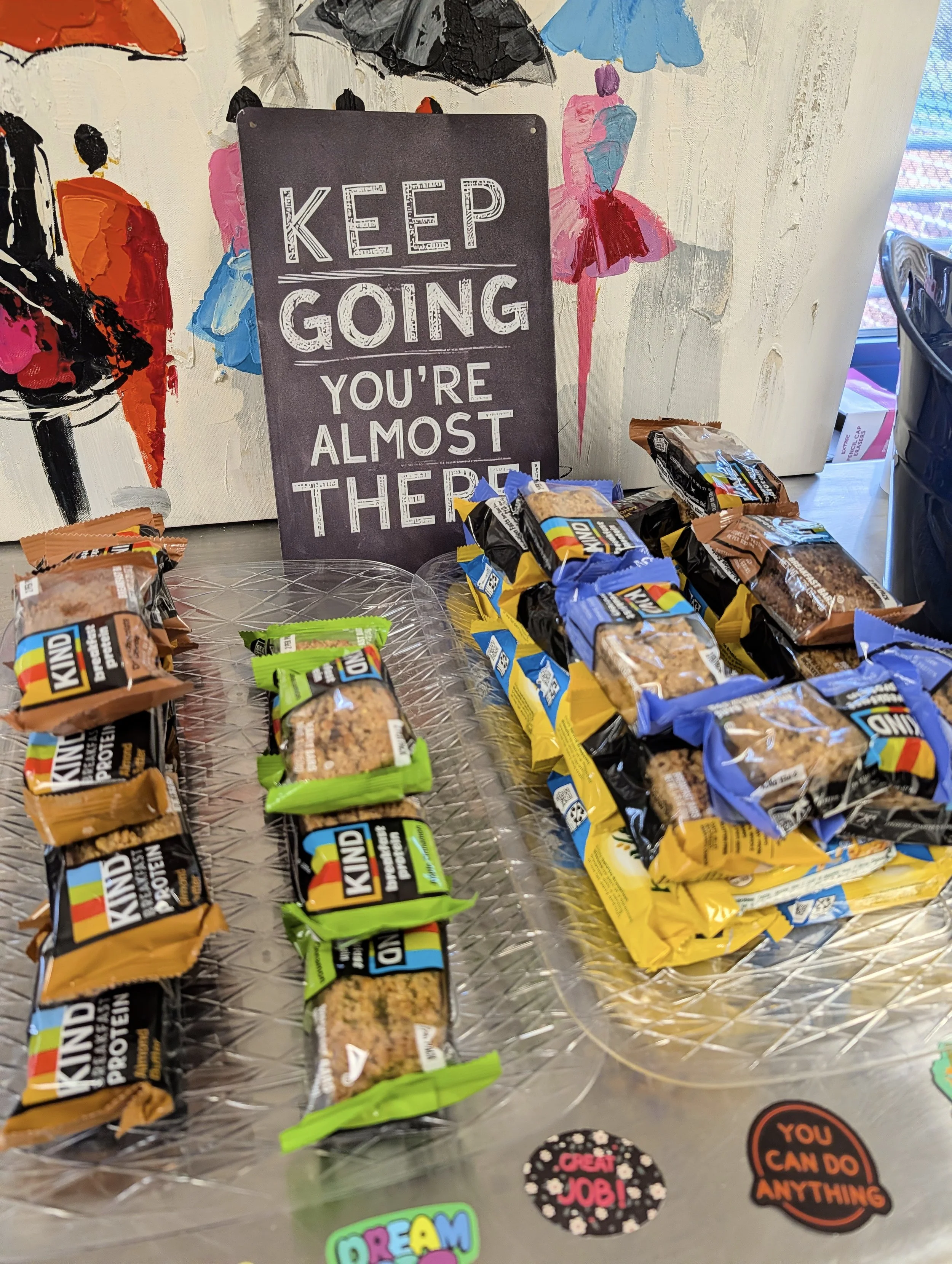 A sign with the motivational message, 'Keep going, you're almost there!' surrounded by various colorful stickers with positive messages and a display of snack bars on a table.