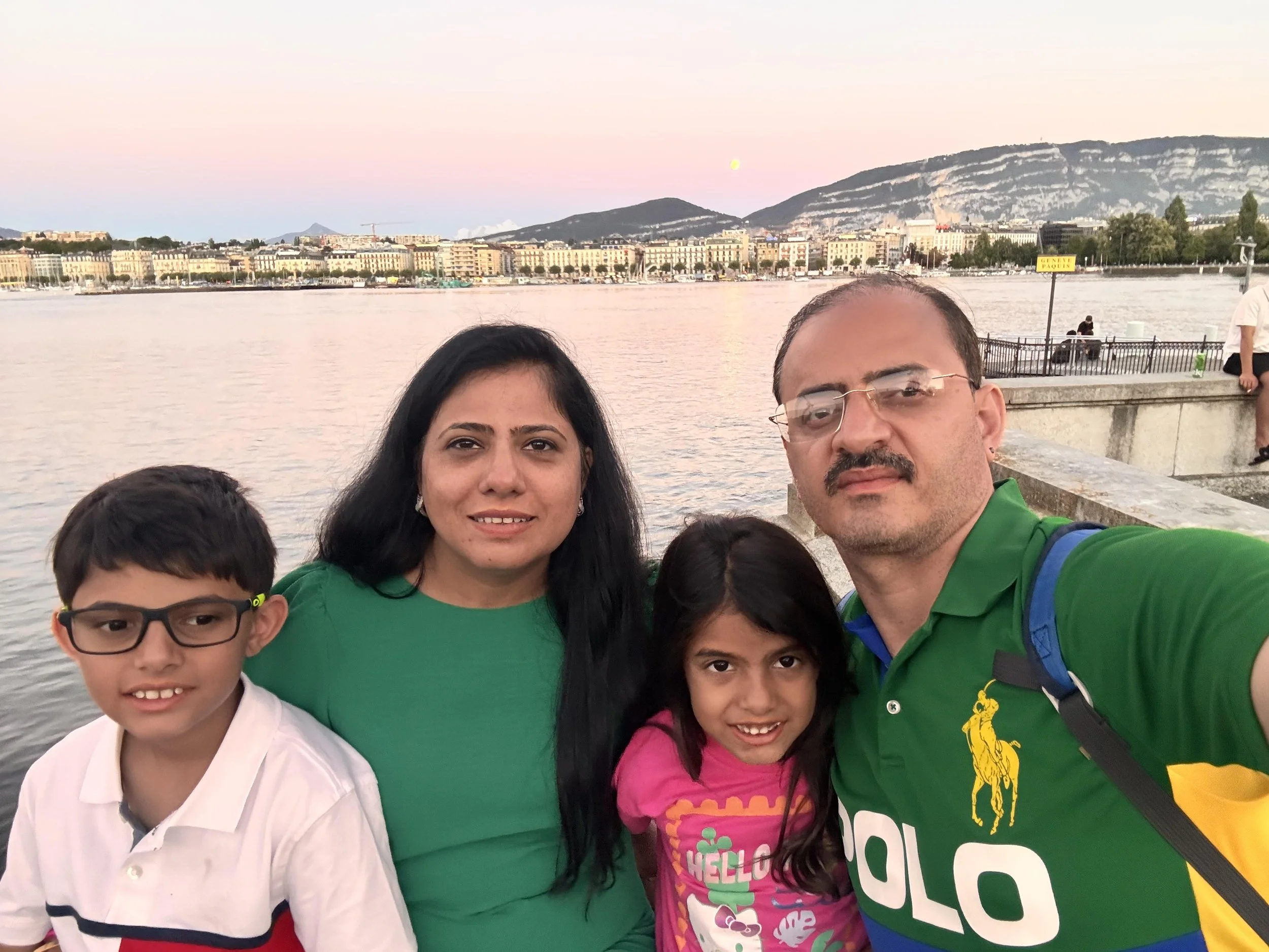 A man, a woman, and two children posing in front of a cityscape backdrop with tall buildings and a mountain landscape. They are smiling and standing on a carpeted floor.