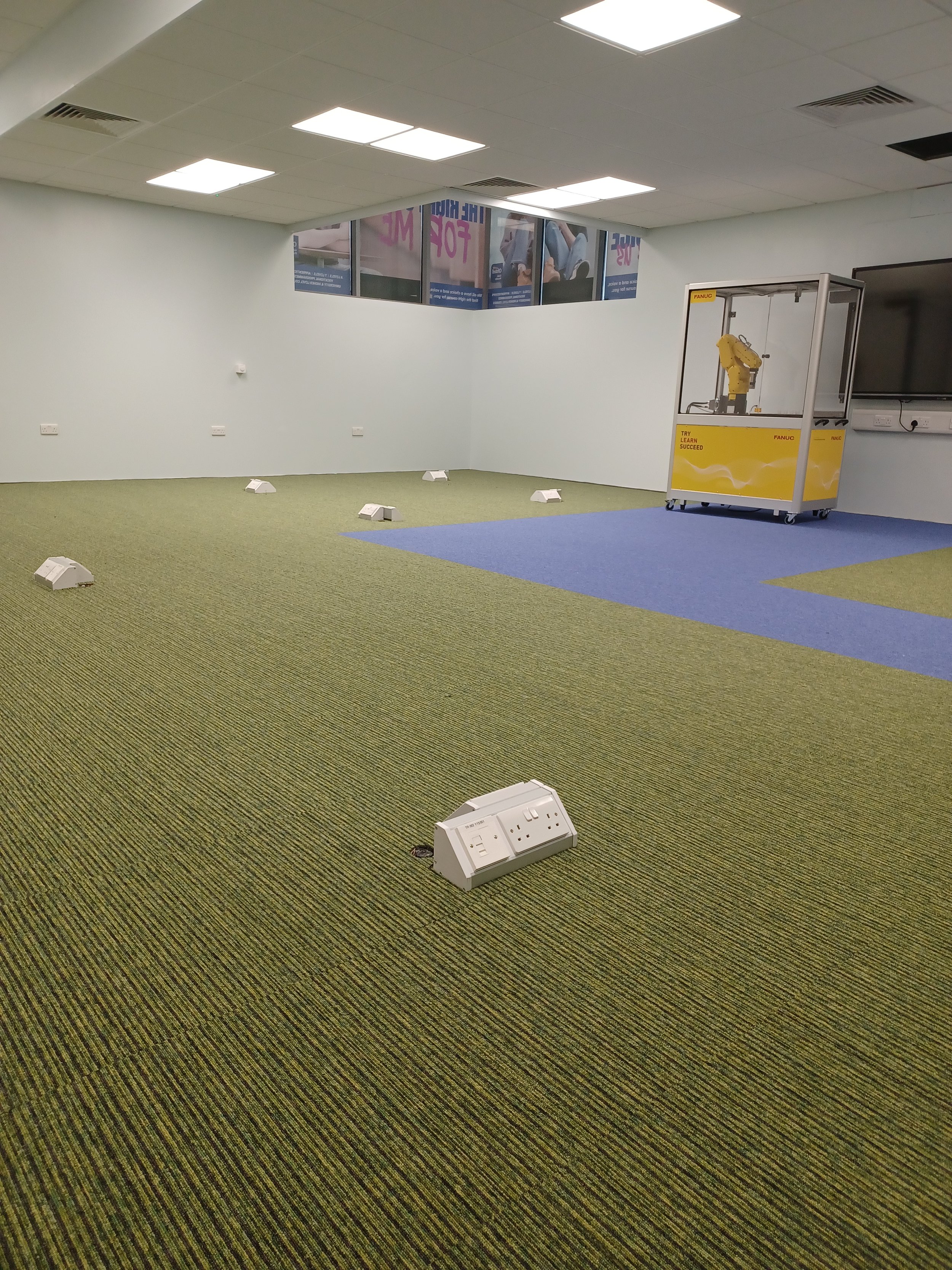 The image shows a university robotics lab with a green carpet tile floor that has a subtle texture. A blue carpet section is laid out in a rectangular shape. Several white equipment units are scattered throughout the space.