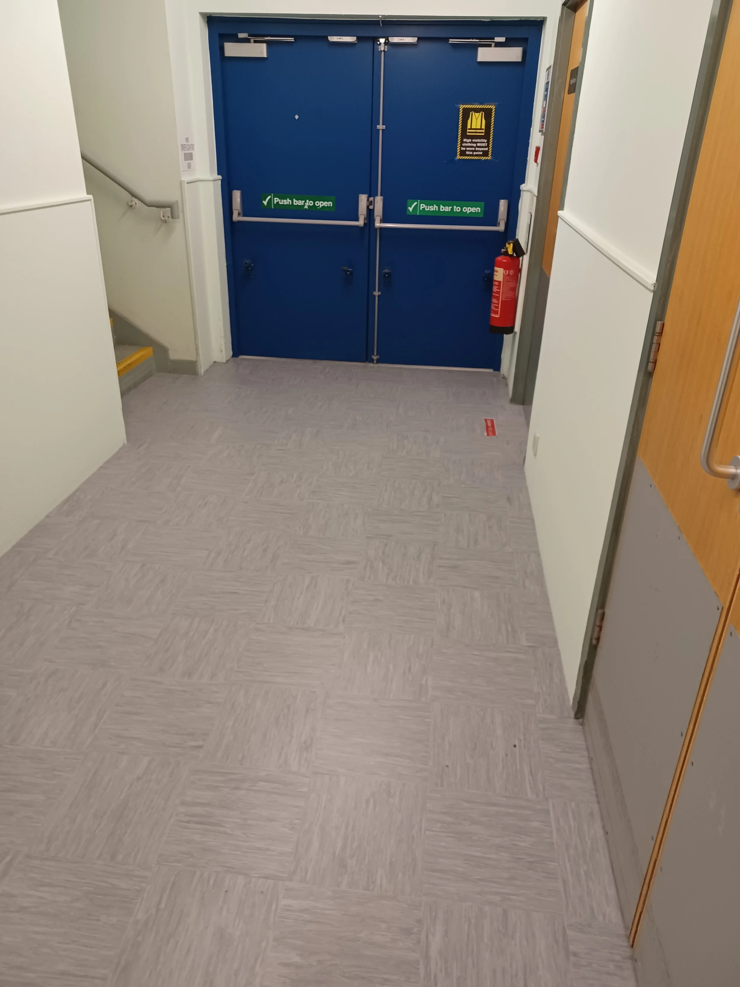 Blue double doors with push bar to open, fire extinguisher on the right, warning signs, and white walls in a hallway with tiled beige flooring.