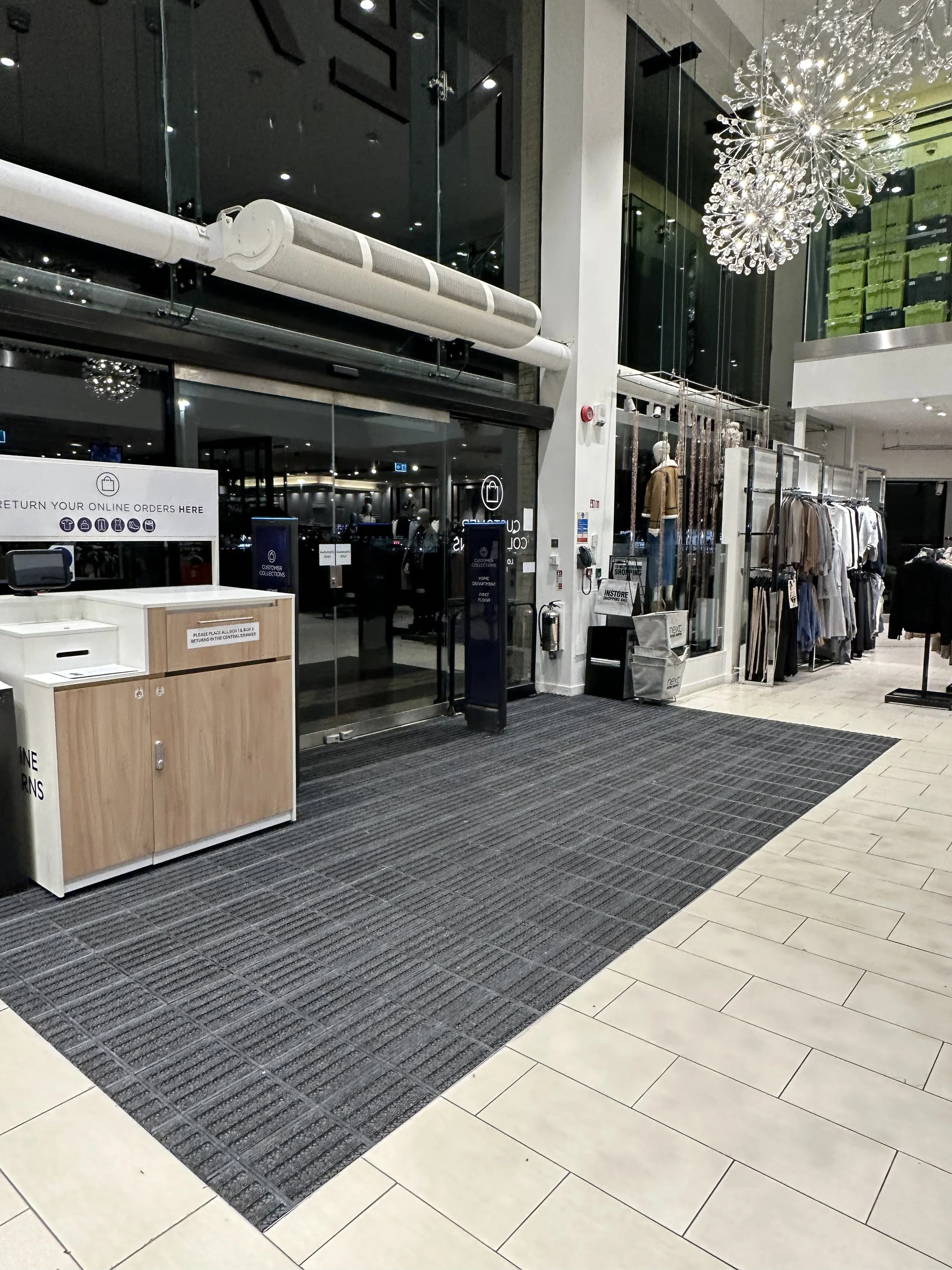 Indoor shopping mall entrance with glass doors, clothing displays on the right, and a checkout counter on the left. The area features a black and white color scheme with tiled flooring and decorative lighting fixtures hanging from the ceiling.