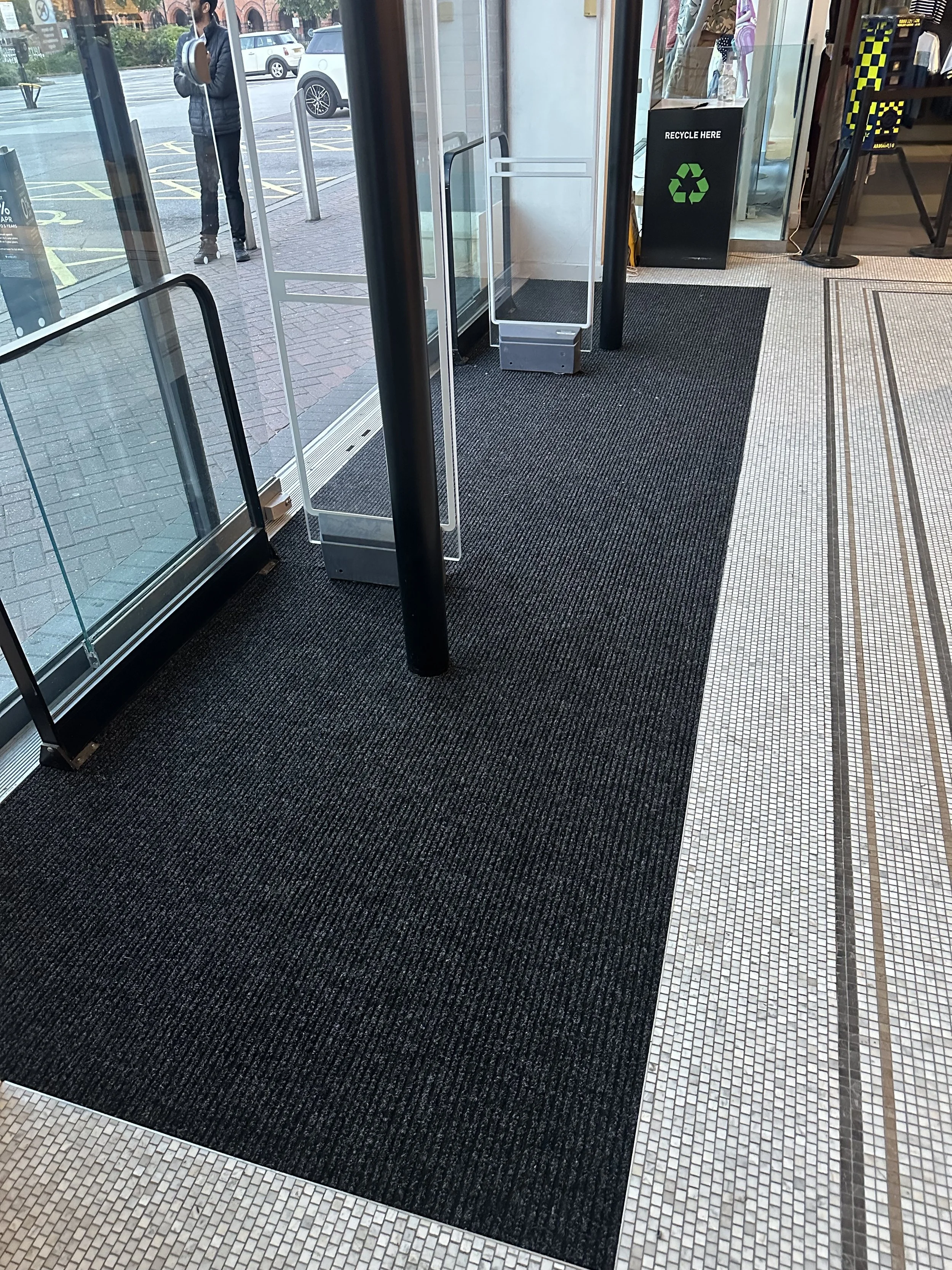 A close-up view of black ribbed entrance matting located in a retail environment. The matting is positioned near an entrance and has been recently replaced due to maintenance.
