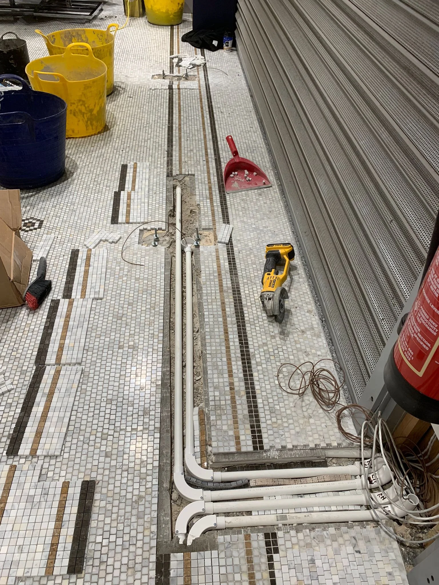 The image shows ceramic mosaic tiles in a retail store with several tiles removed to expose underfloor pipework. The tiles are partially lifted, and plastic piping runs along the exposed area, indicating maintenance work is in progress. 