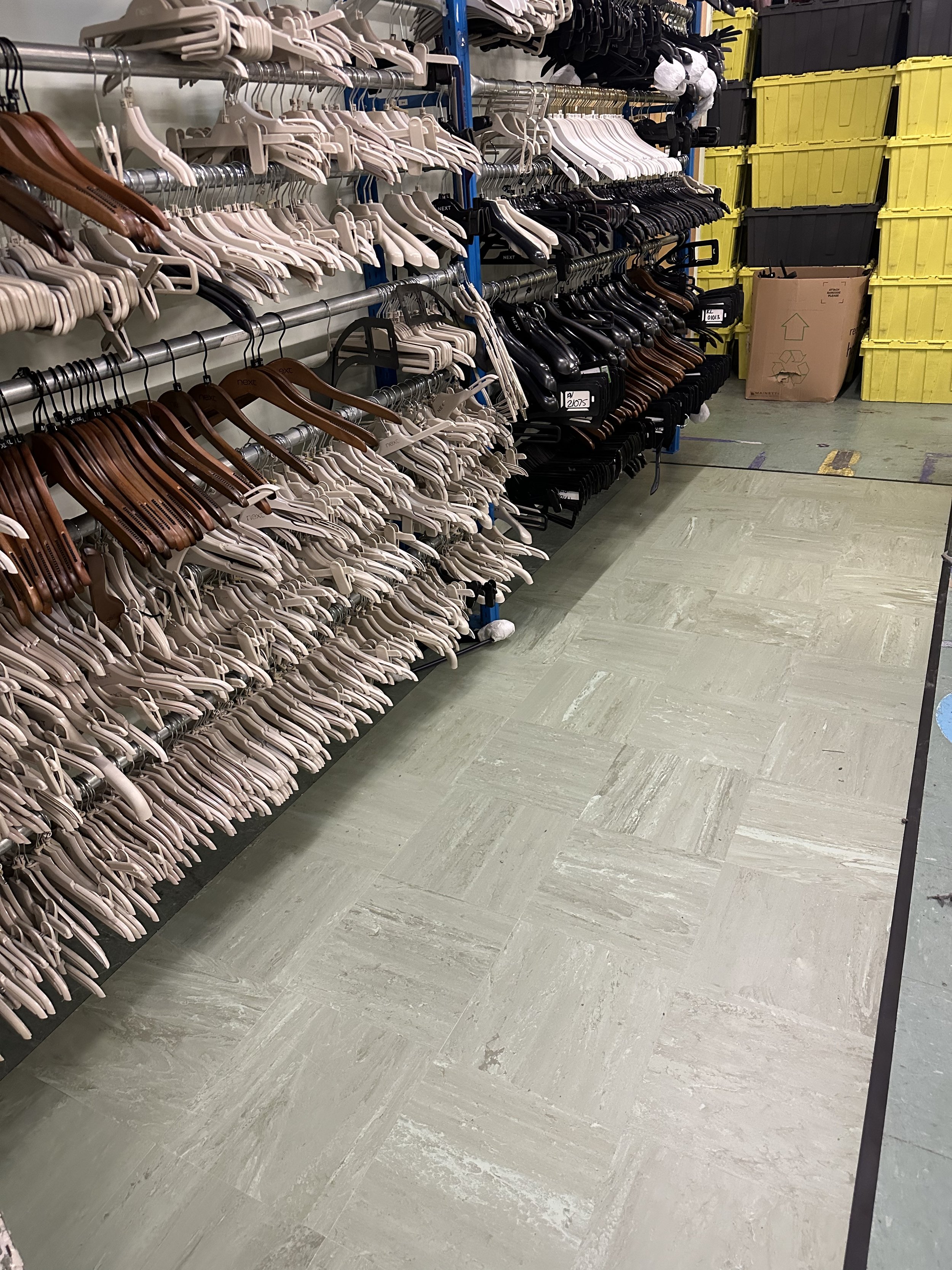 A stockroom showing vinyl tile flooring that appears recently installed. Rows of clothing hangers are neatly arranged on racks to the left, including wooden and plastic styles. The floor is a pale green colour with a subtle texture.