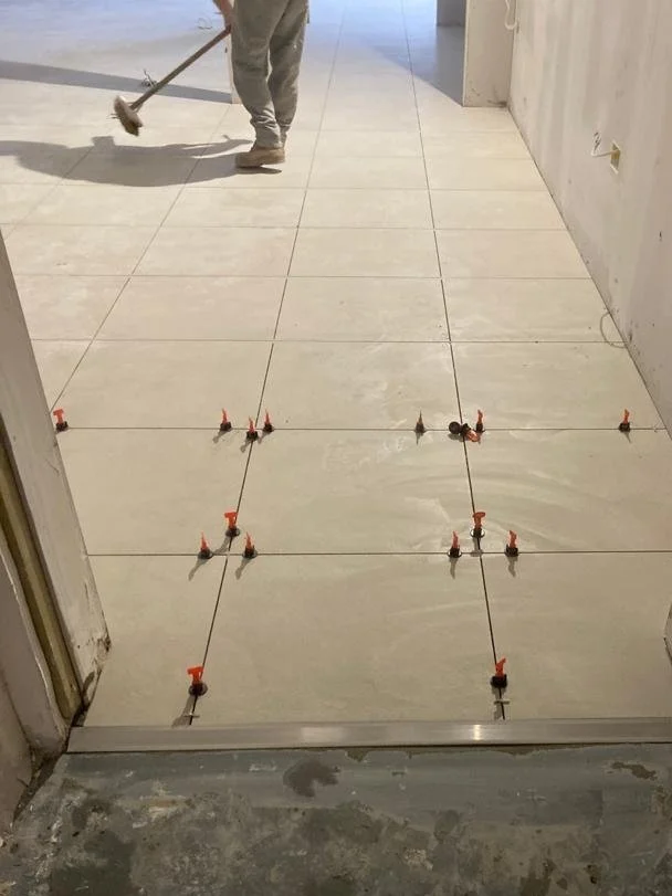 The image shows newly installed beige ceramic tiles in a lift. Spacers are positioned between the tiles, indicating they are ready for grouting. The old cracked tiles have been replaced.