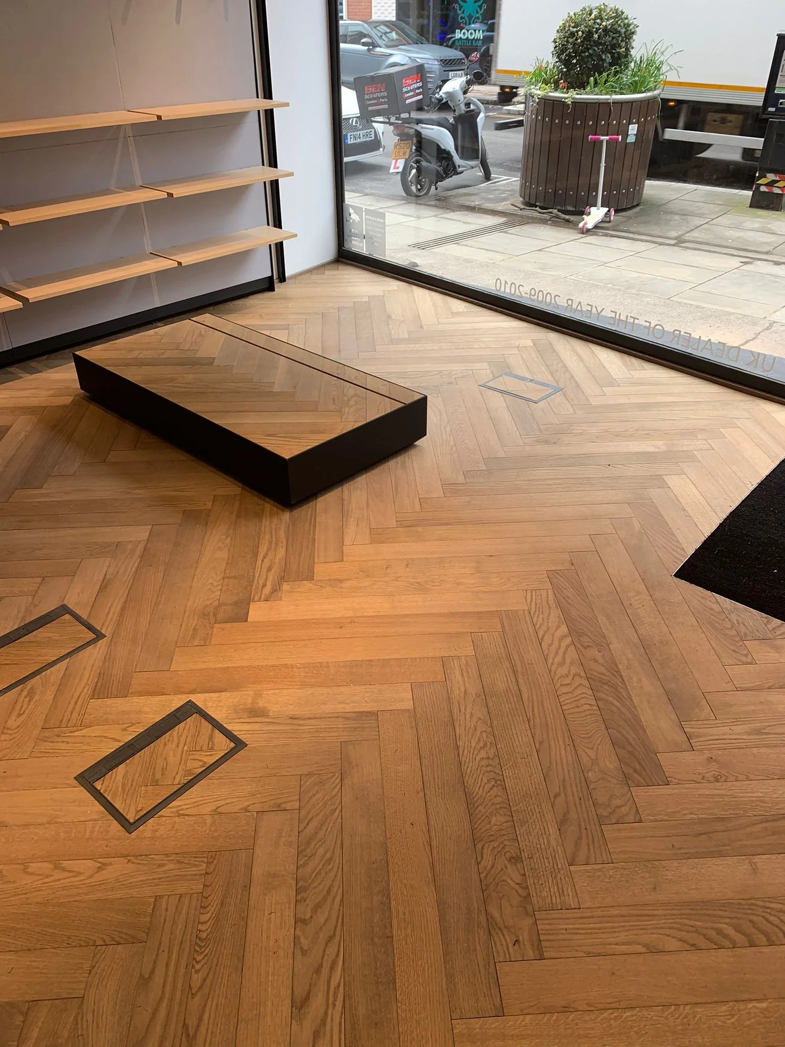 Empty interior of a bookstore or shop with wooden herringbone flooring, a black display table, empty wooden wall shelves, and a large window showing a street view with a scooter, a large planter with a bush, and a pink scooter outside.