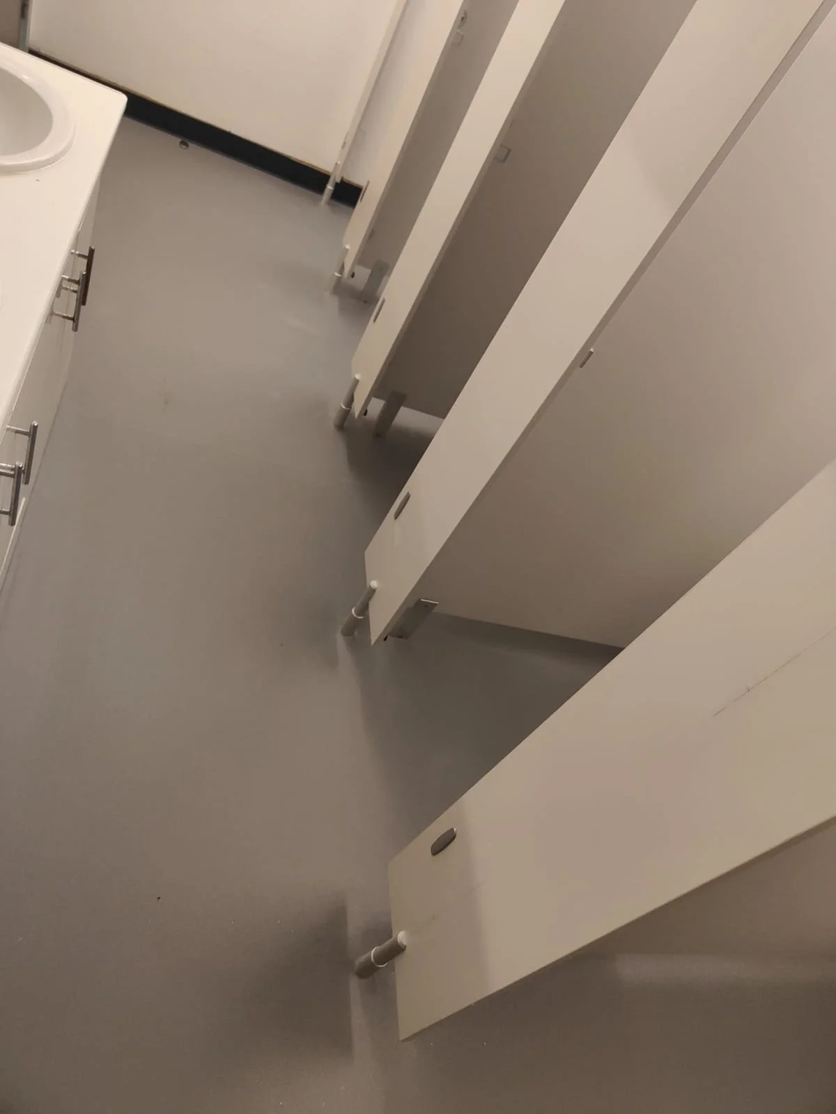 The image shows the interior of a toilet with newly installed sheet vinyl flooring. Light grey flooring extends across the space, whilst white partition walls separate toilet cubicles, creating a clean and modern appearance.