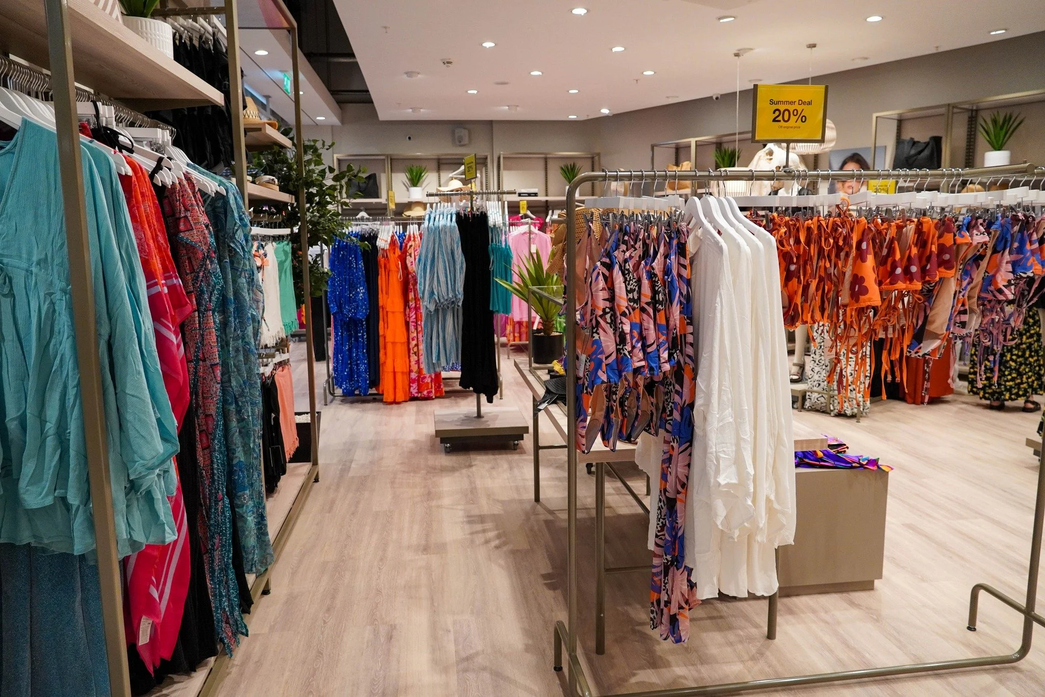 Interior of a clothing store with racks of colorful dresses and outfits, some having floral and patterned designs. There are yellow sale signs indicating discounts, and potted plants are placed on shelves in the background.