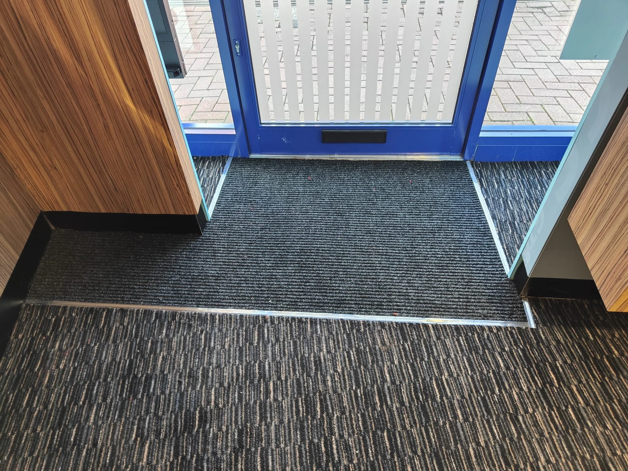 The image captures an irregularly shaped black ribbed matting positioned at the threshold of a shopfront. The matting consists of multiple horizontal ridges which enhance traction, ideal for capturing dirt and moisture.