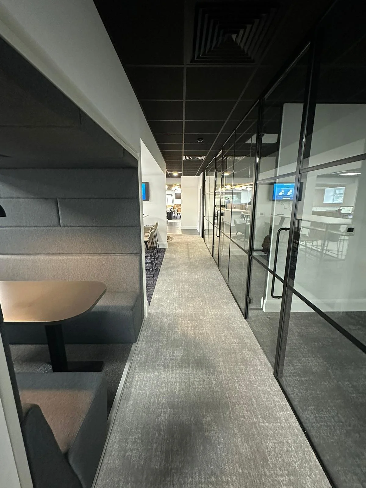 The image shows a contemporary office interior focusing on a central hallway with light grey carpet tiles. The hallway is flanked by glass-walled meeting rooms, allowing visibility of the space beyond.