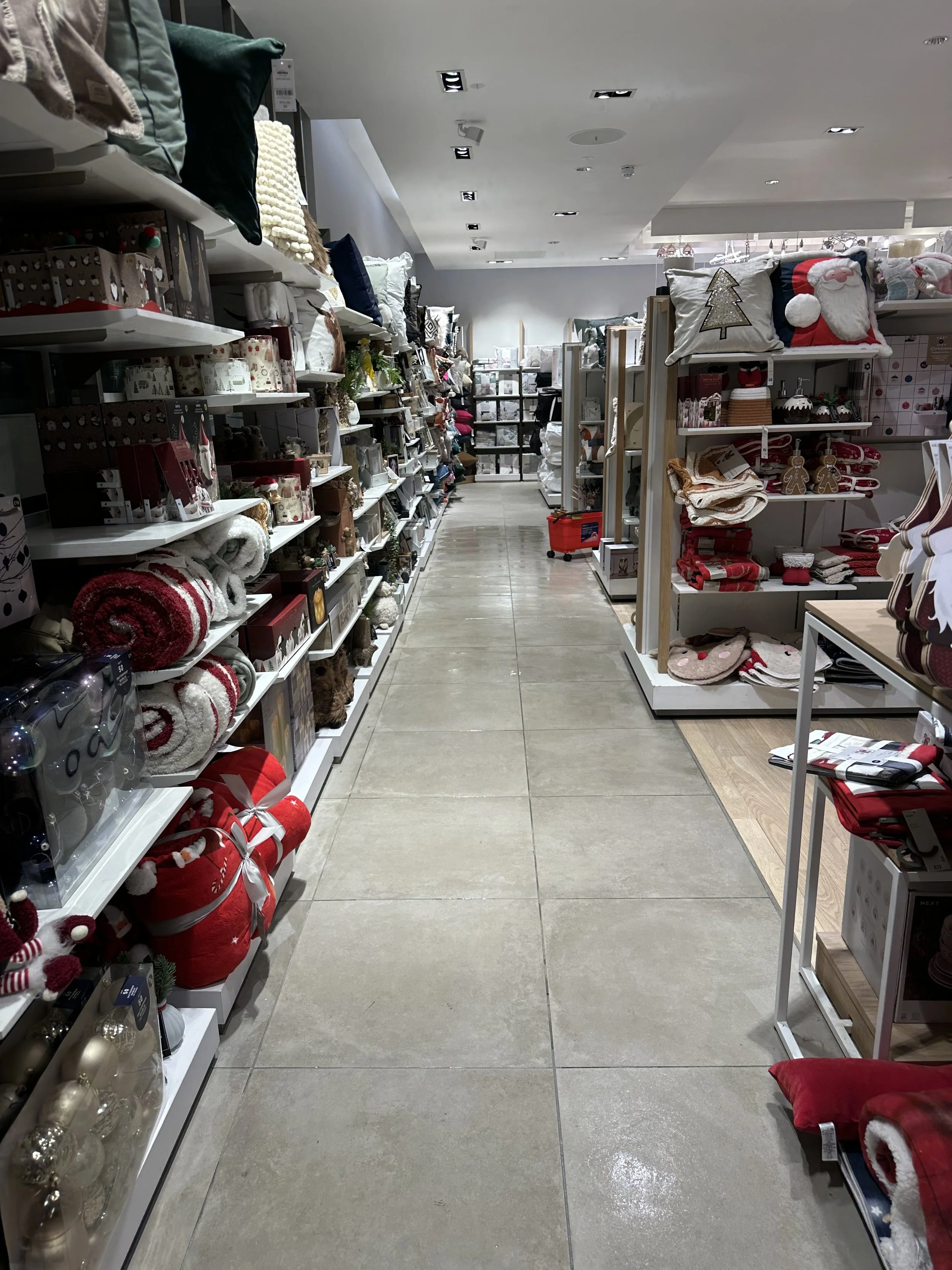 Inside a retail store, the image shows an aisle where merchandise is displayed on both sides of the shelves. The flooring is ceramic tiles that have recently been installed and grouted, resulting in a partially wet appearance.