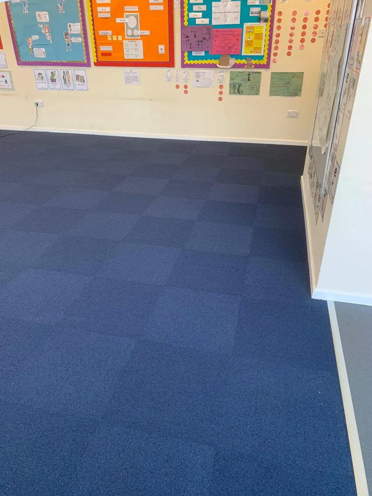 The image displays blue carpet tile flooring in a classroom, with square tiles arranged in a grid layout. The texture of the tiles is visible, providing a soft and durable surface. 