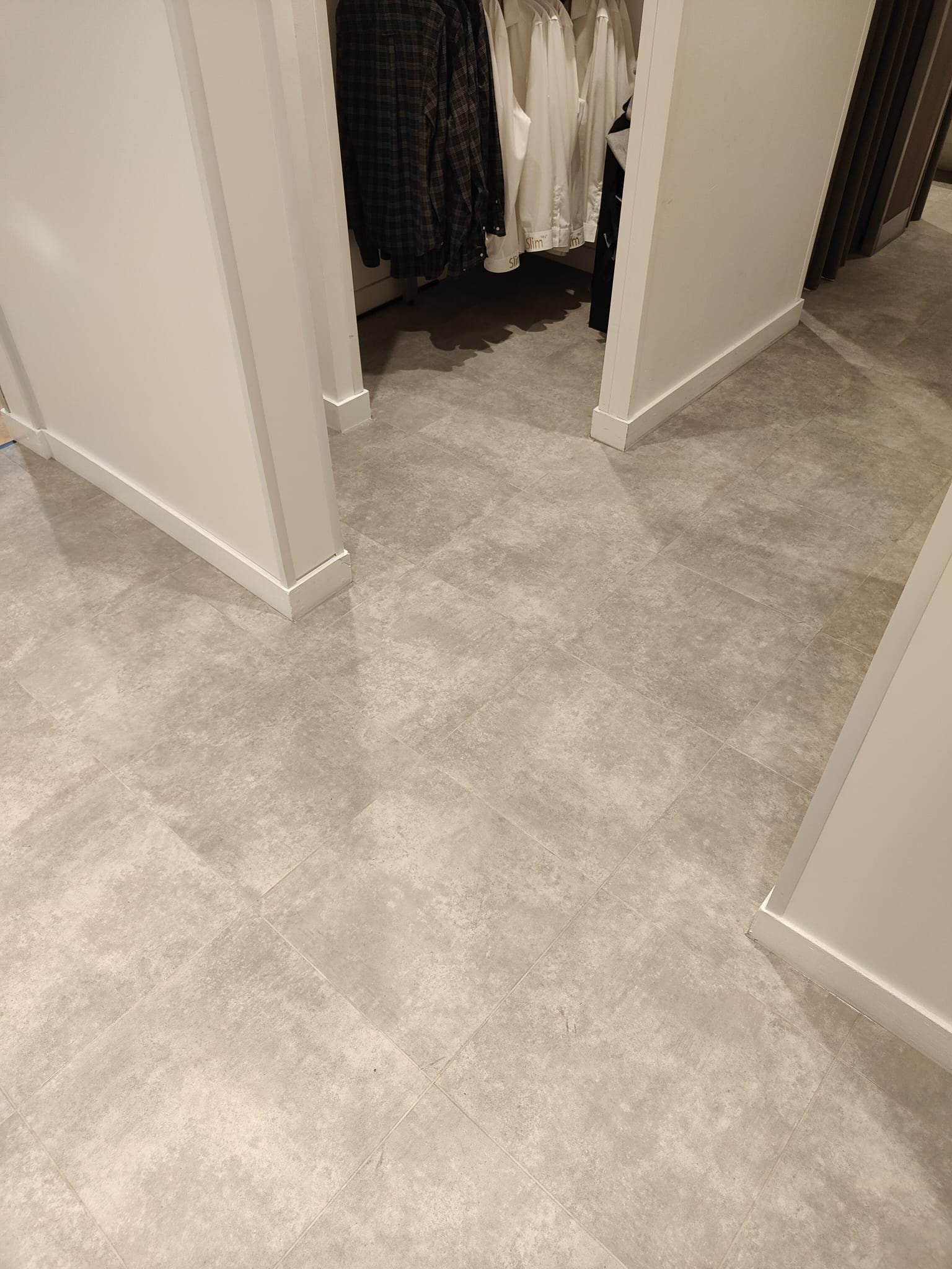 The image shows grey luxury vinyl tile (LVT) flooring laid in a square pattern, situated in the lobby area of a retail changing room. The floor appears clean and well-maintained, with light reflecting off its smooth surface. 