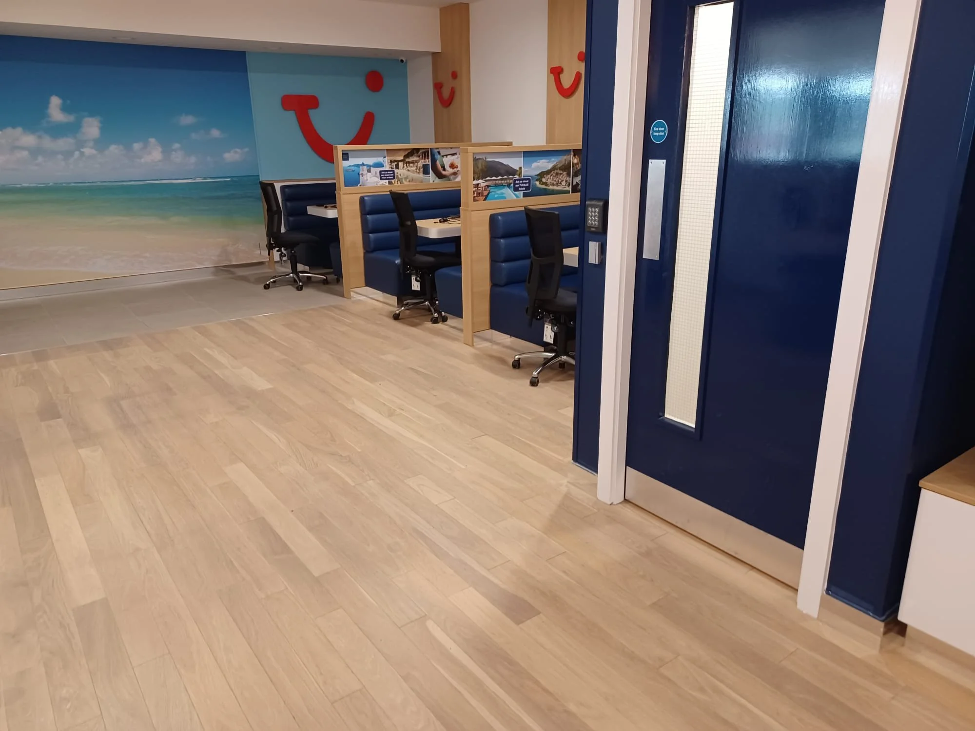 The image showcases a travel agency's interior with a light wood LVT flooring installation. Three seating booths, upholstered in a deep blue, line the back wall.