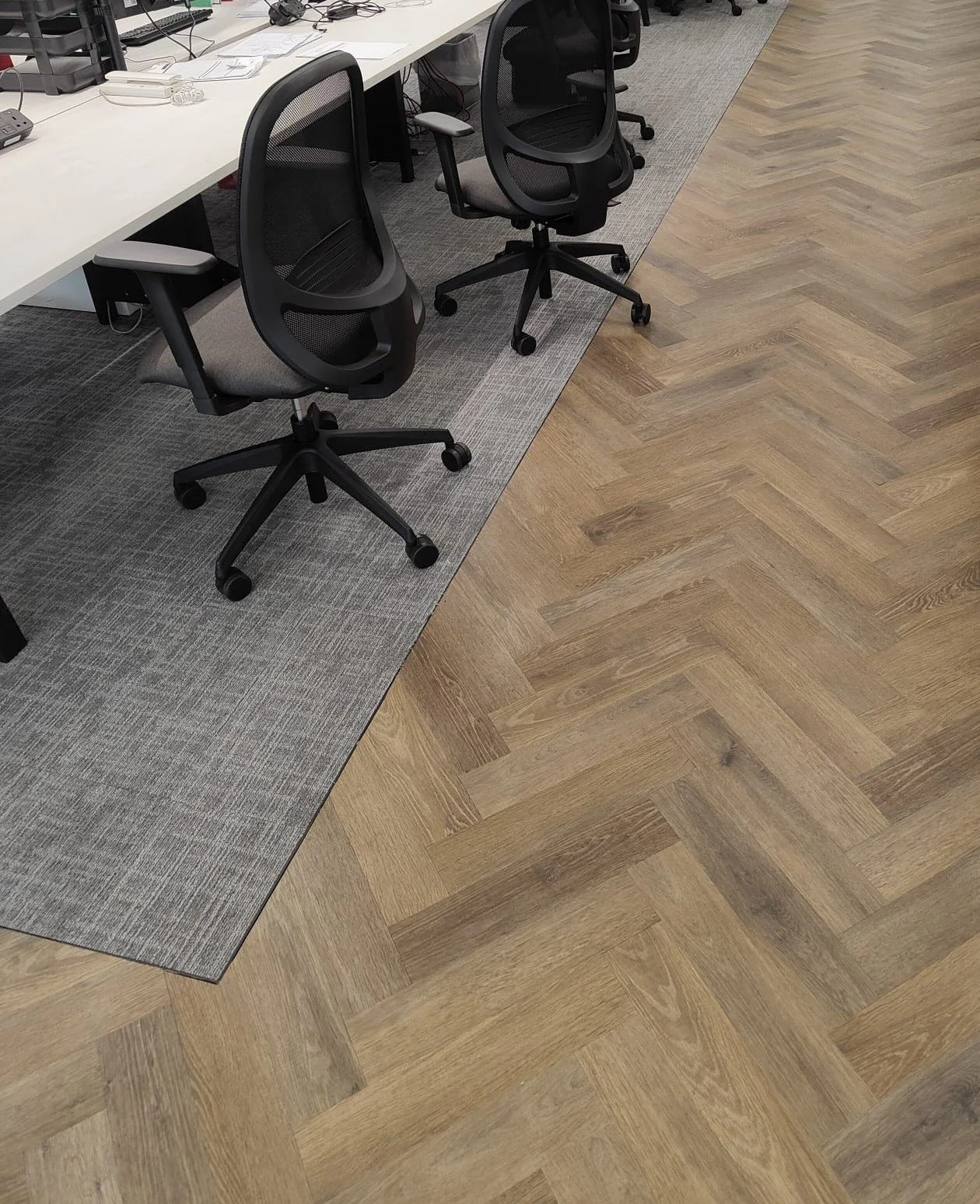 Office workspace with white desks, black ergonomic chairs, and a mix of carpet and wooden flooring.