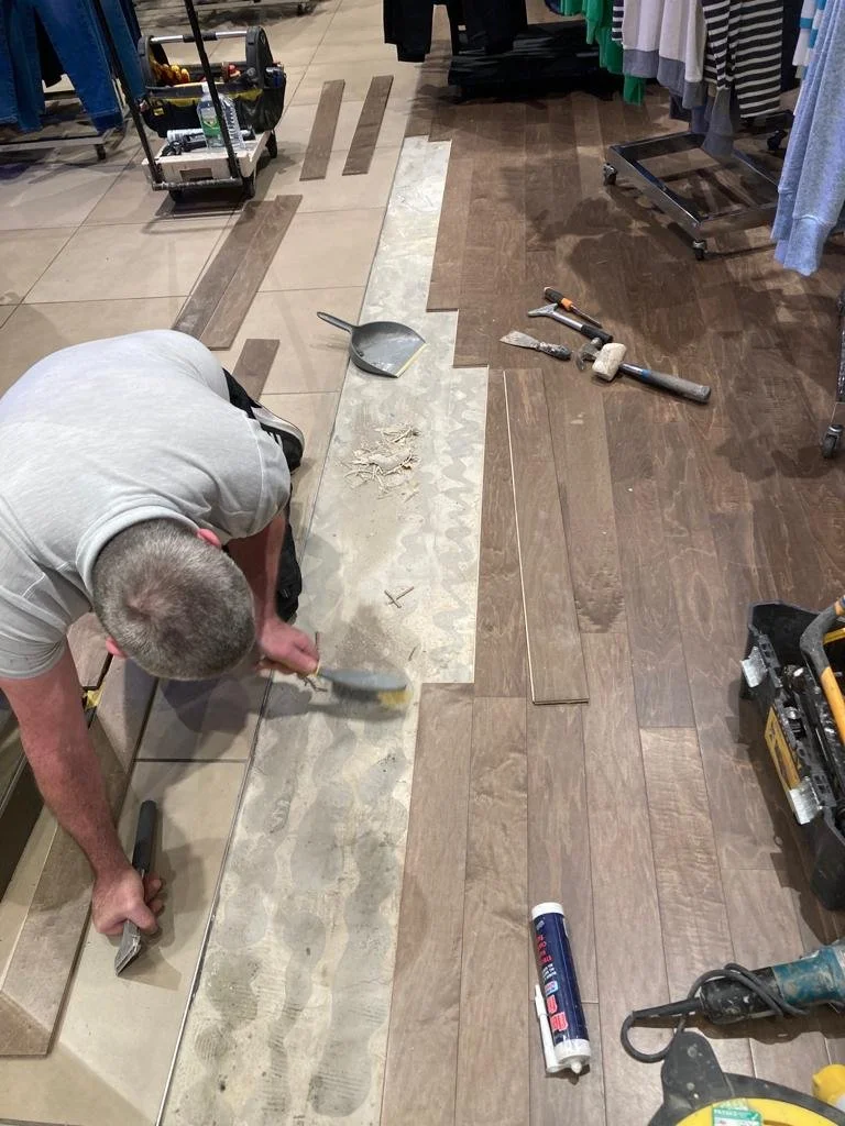 An image showing the fitter using a brush to remove dust and debris from the floor. He has uplifted some of the timber flooring planks, his tools are lay on the floor beside him.