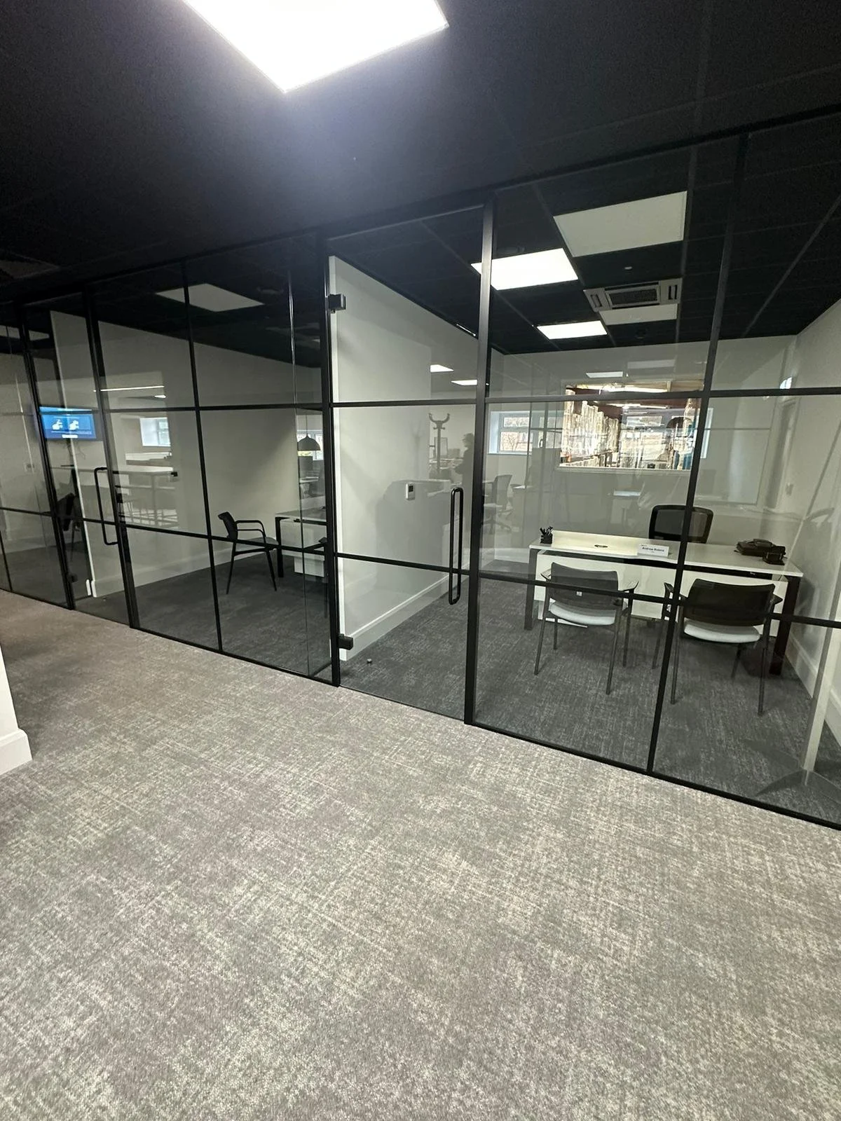 The composition features a rectangular office area defined by black-framed glass walls. The carpet tile flooring is a light grey, contrasting with the darker chairs. The desk is centrally positioned with a white finish