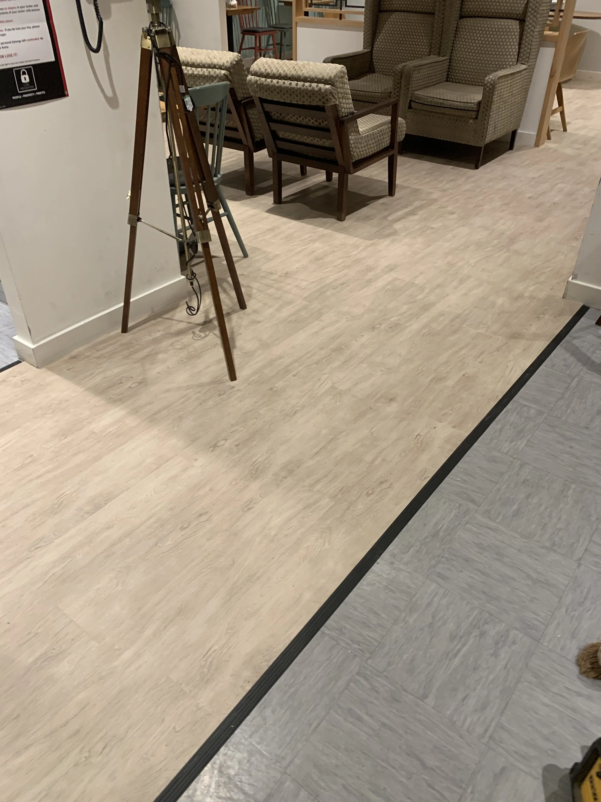 A section of LVT flooring installed in a furniture department. The flooring consists of light wood-like planks with subtle grains, extending across the space. A wooden table with slender legs is partially in view.