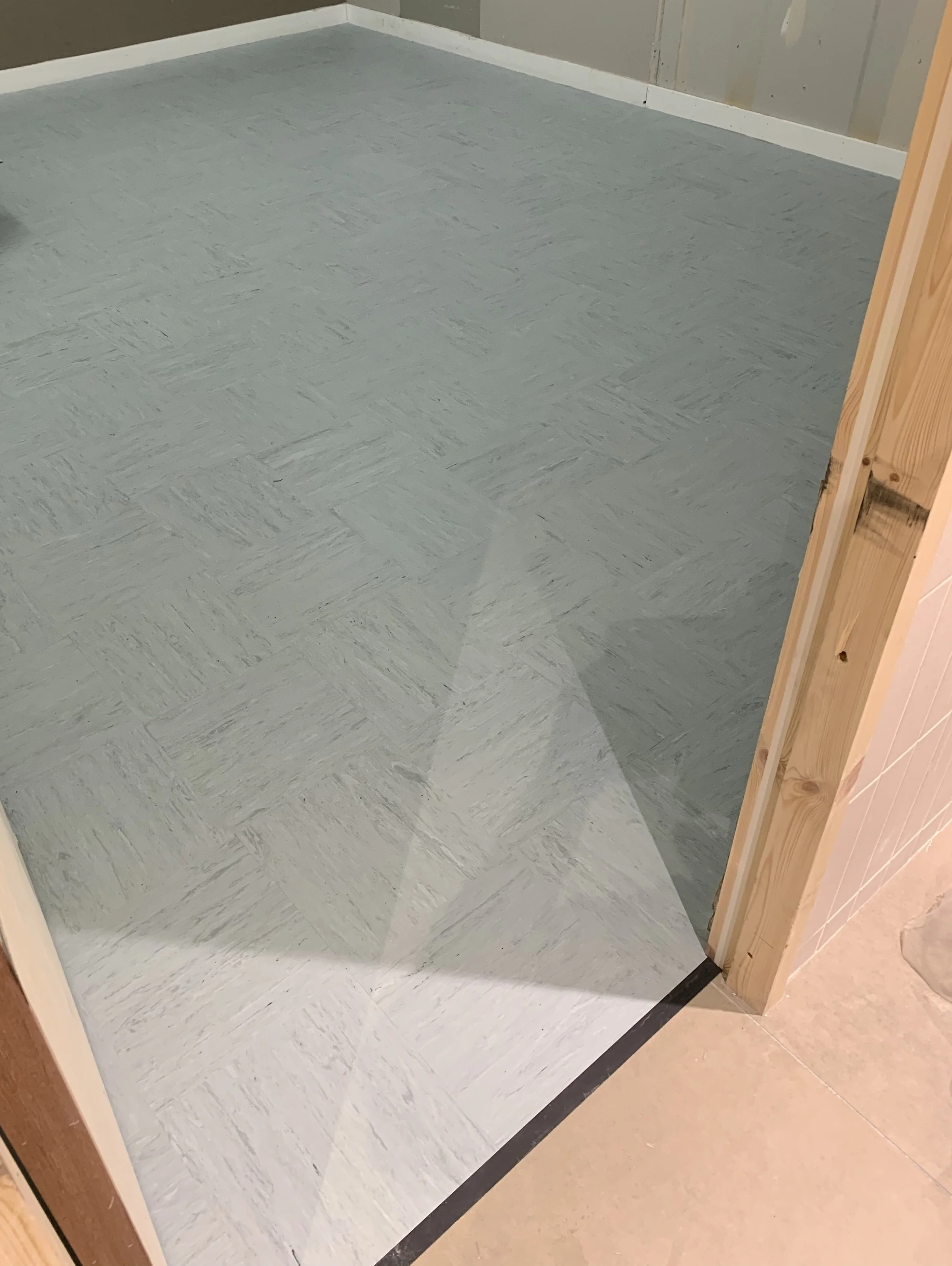 The image shows a section of light grey vinyl tile flooring in a BOH room. The tiles are laid in a square pattern, highlighting the surface's texture and slight variations in colour. A wooden door frame visible at the edge of the image.