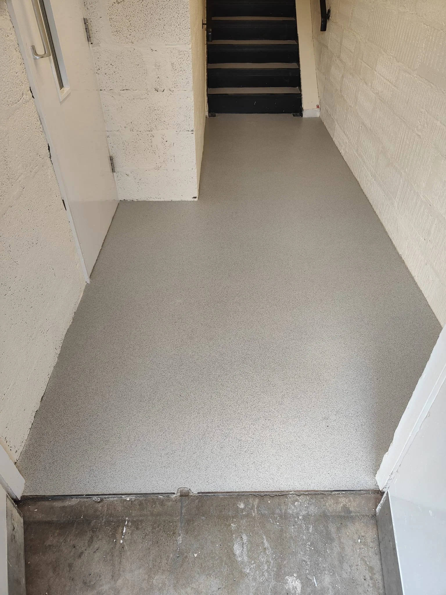 The image shows a newly laid sheet vinyl floor, predominantly light grey with a subtle speckled texture. The flooring covers the entire visible area, seamlessly connecting to a plain wall on the left, which has a rough, unfinished surface. 