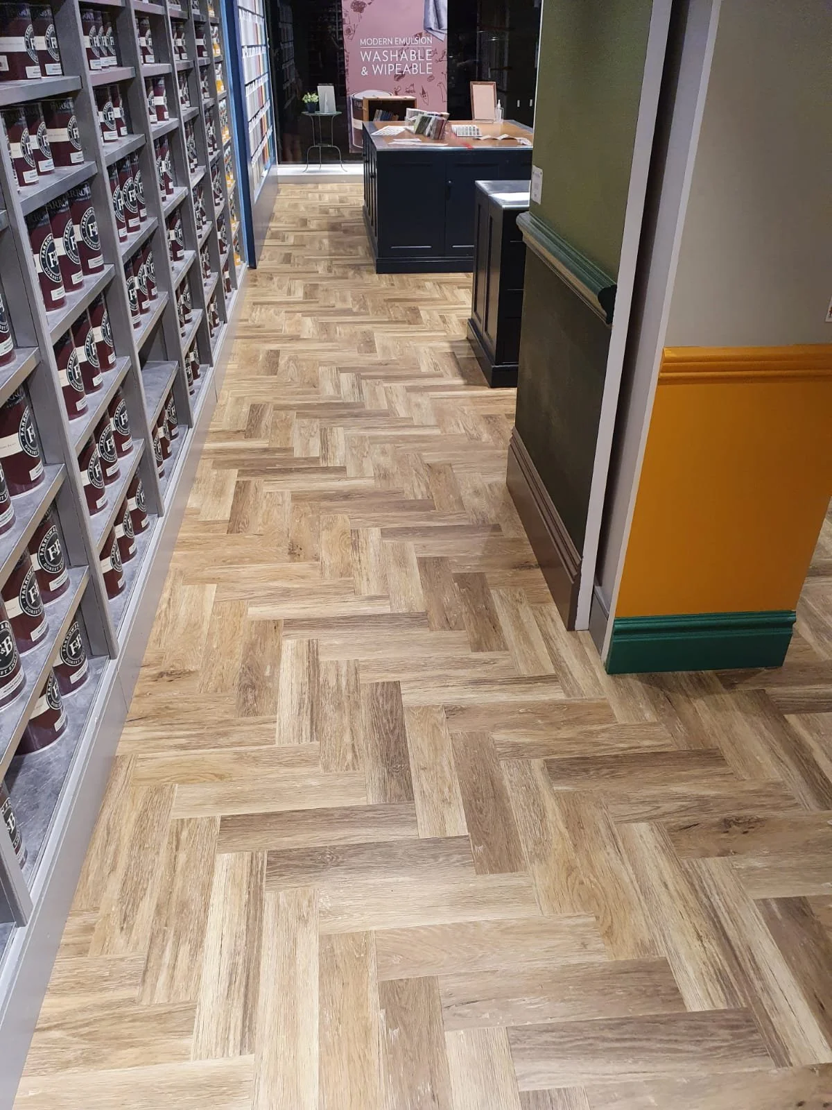 The image shows a herringbone-patterned LVT wood floor located in a store aisle. The floor is made of varying shades of brown and cream. On the left side, shelves display tinned paint, whilst a colourful wall is visible on the right.