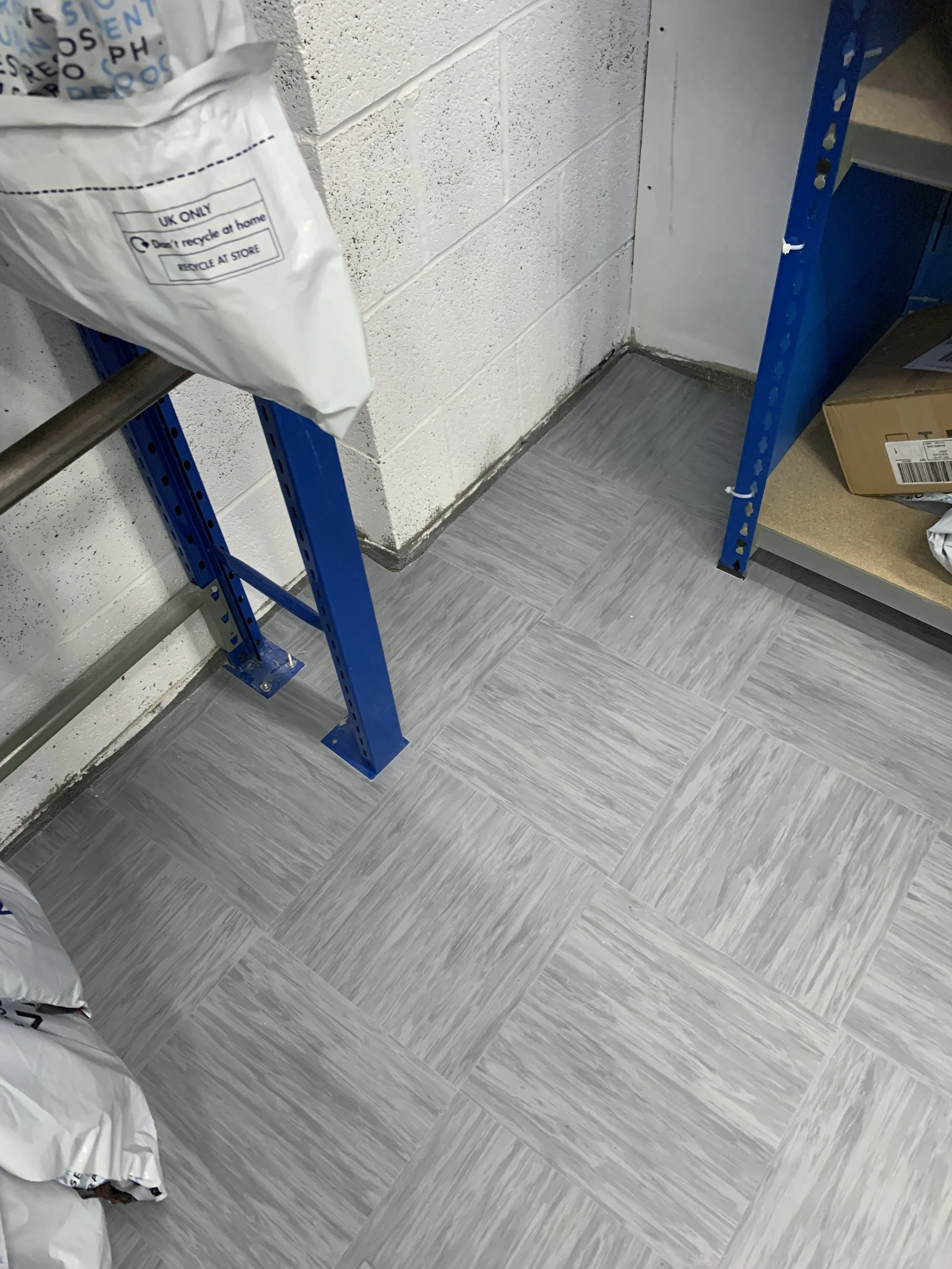 The image captures a corner of a stockroom featuring newly installed light grey vinyl tile flooring. The tiles are arranged in a square pattern with a subtle texture.
