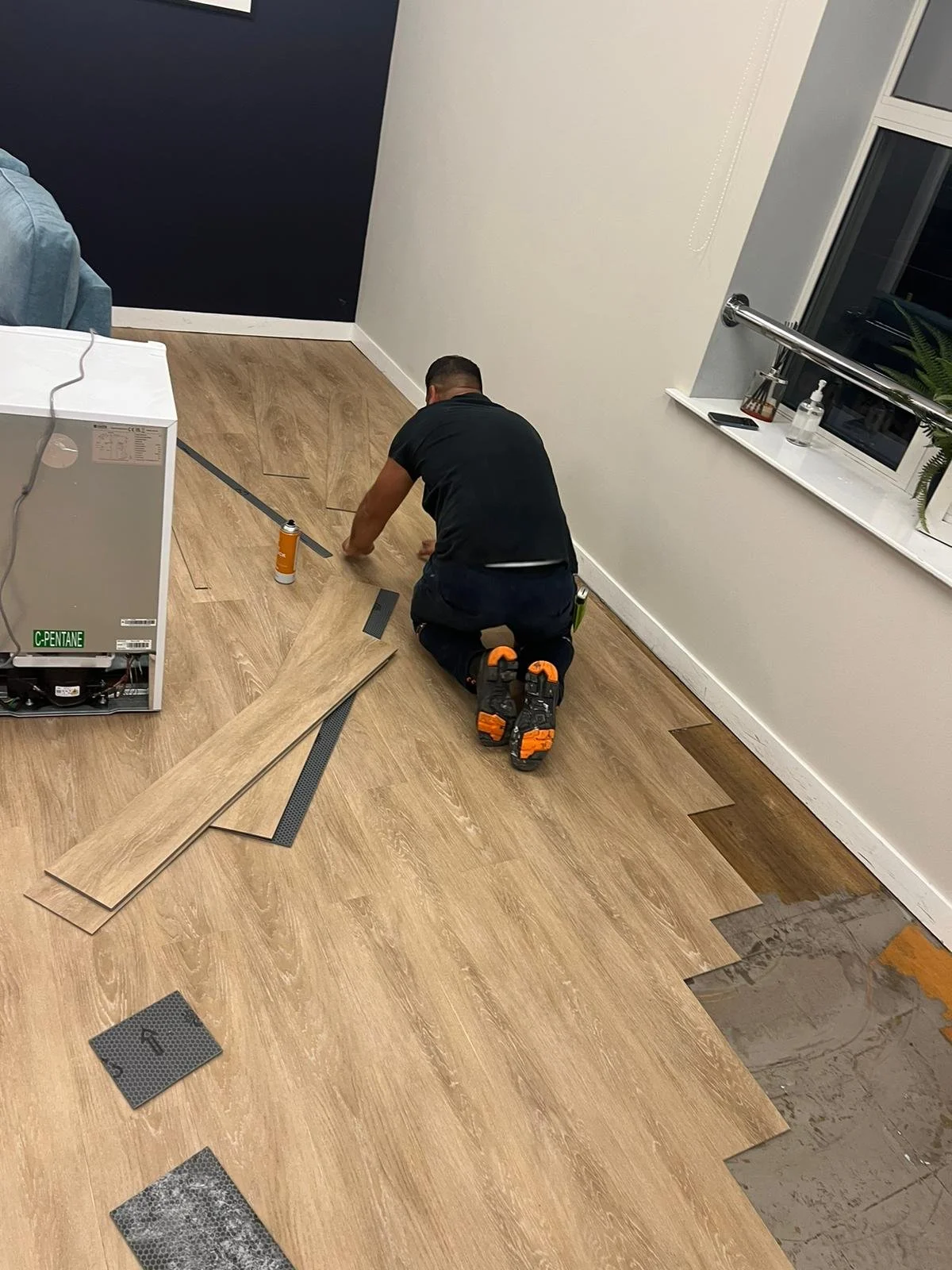 A flooring fitter is on his knees while installing LVT planks in a staffroom area. Loose laid planks are arranged on the floor.