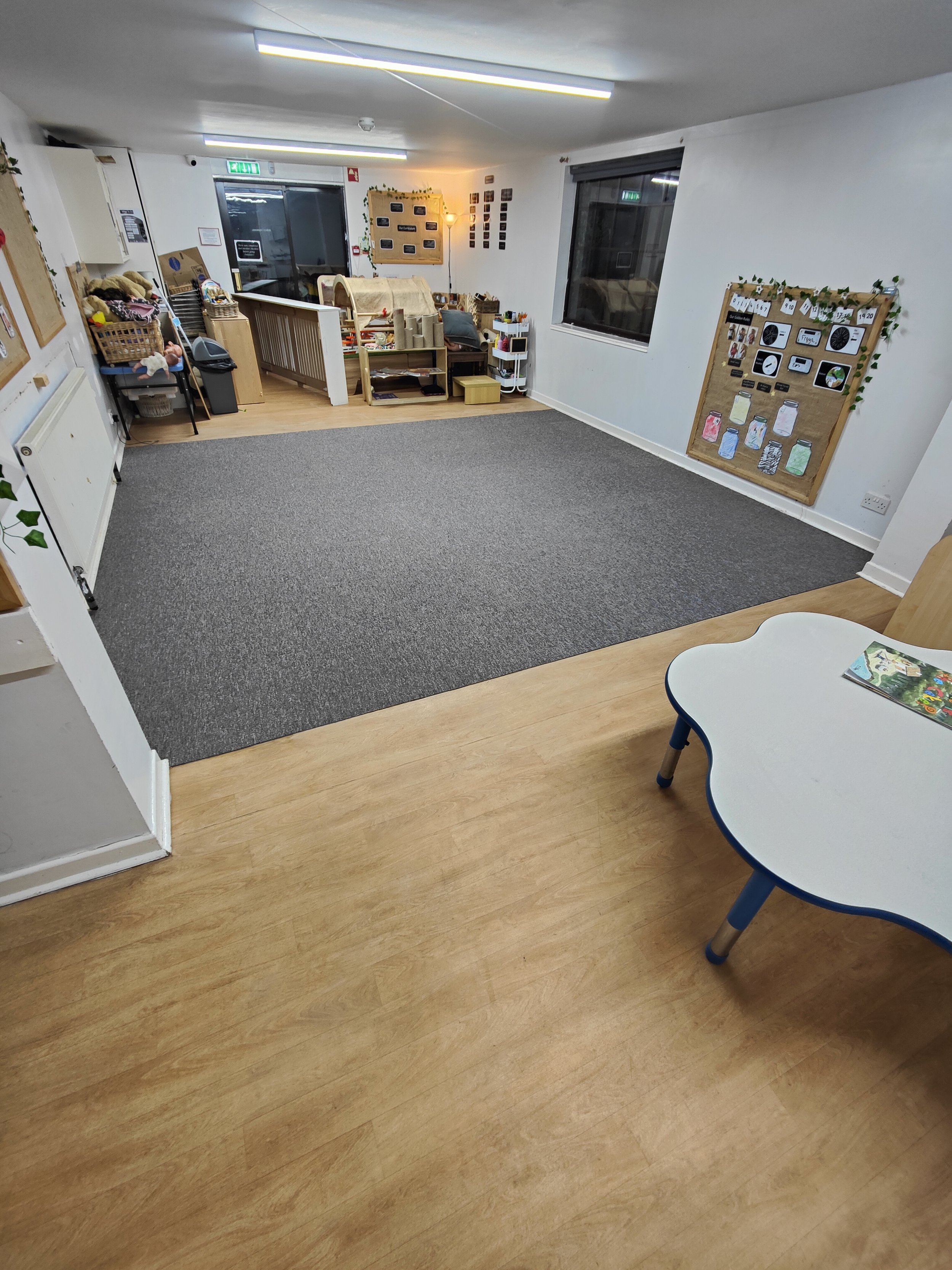 The image features a nursery with a surface of dark grey carpet tiles, covering a significant area of the floor. The flooring juxtaposes with wood-coloured LVT in the foreground. A white children's table is in the image's lower right corner