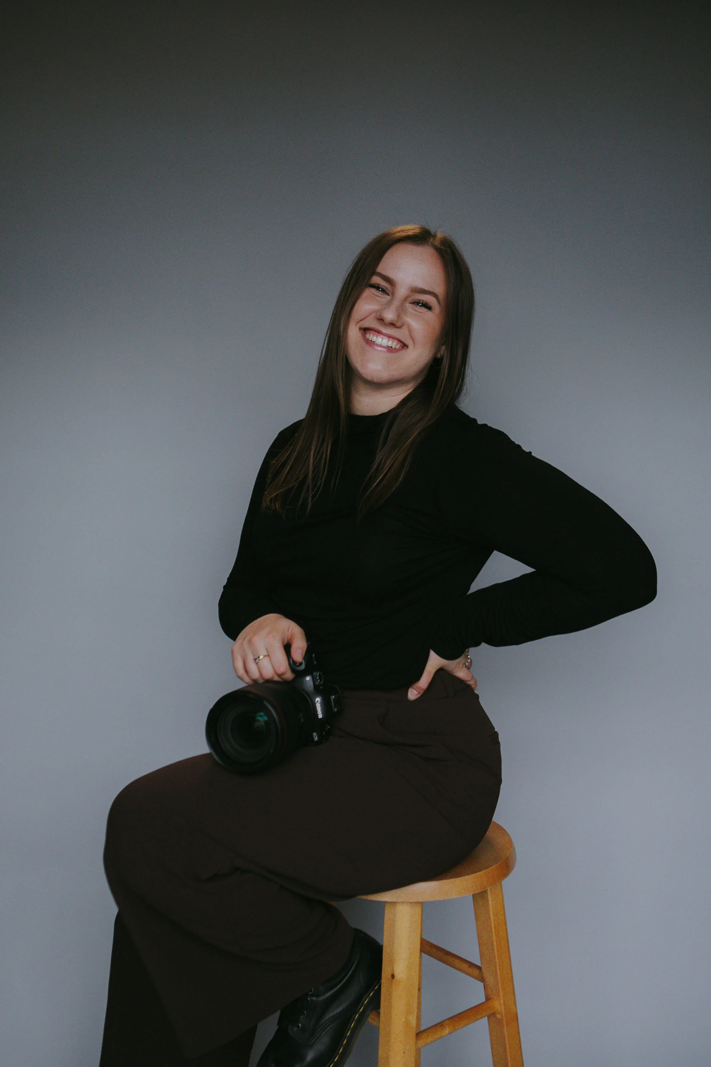 Portrait of a woman holding a camera wearing all black sitting on a stool with a grey background