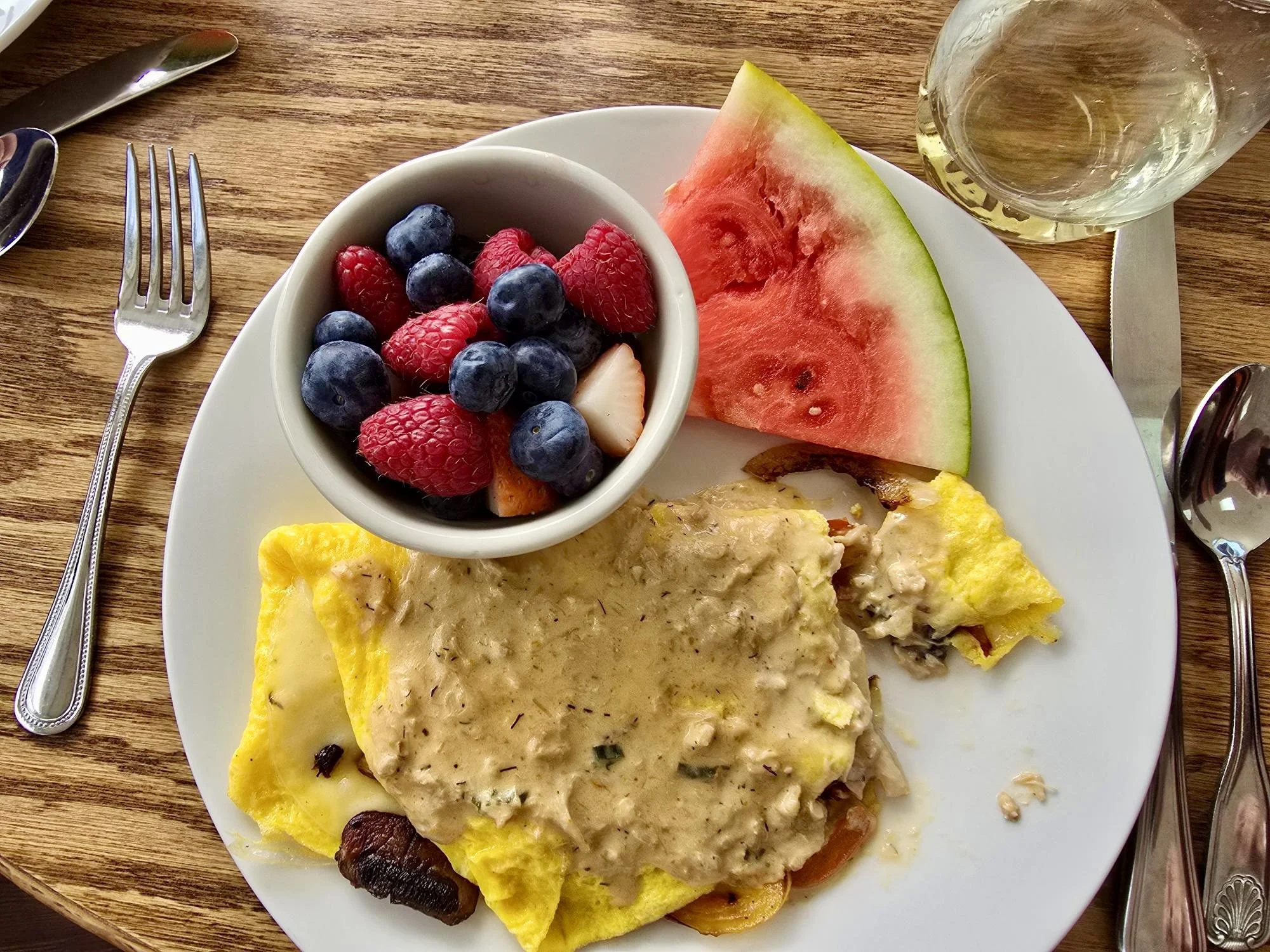 Two eggs Benedict on a plate with microgreens, topped with melted cheese, in a restaurant with a lake view, a cocktail glass, and other drinks in the background.
