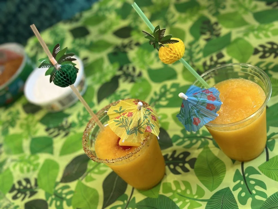 Two tropical drinks with colorful paper umbrellas and fruity garnishes on a green leafy tablecloth.