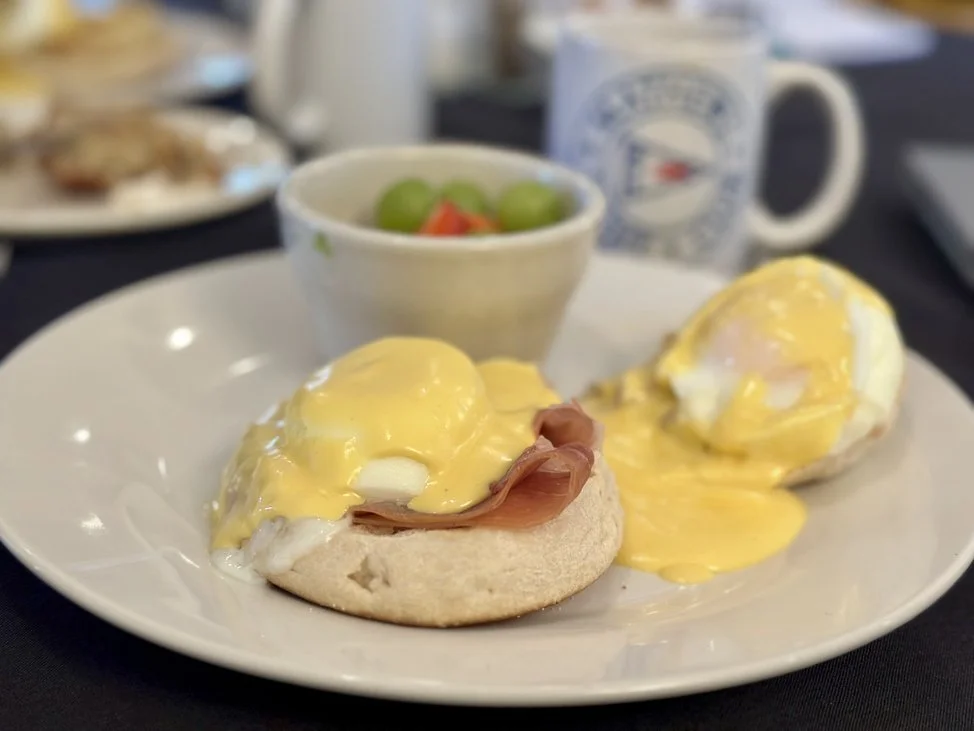 Two eggs Benedict with ham on a toasted English muffin, hollandaise sauce, a small bowl of green grapes and strawberries, and a mug in the background.