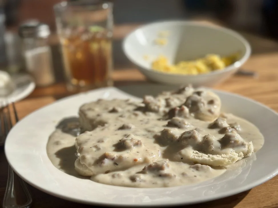 A plate of biscuits with sausage gravy on top, with a bowl of scrambled eggs and a glass of iced tea in the background.