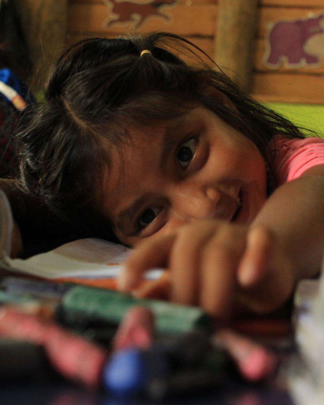 Niña con cabello castaño oscuro y ojo claro, acostada en una mesa, sonriendo, rodeada de tizas de colores y un cuaderno, en un ambiente escolar con paredes de madera y decoraciones de animales.