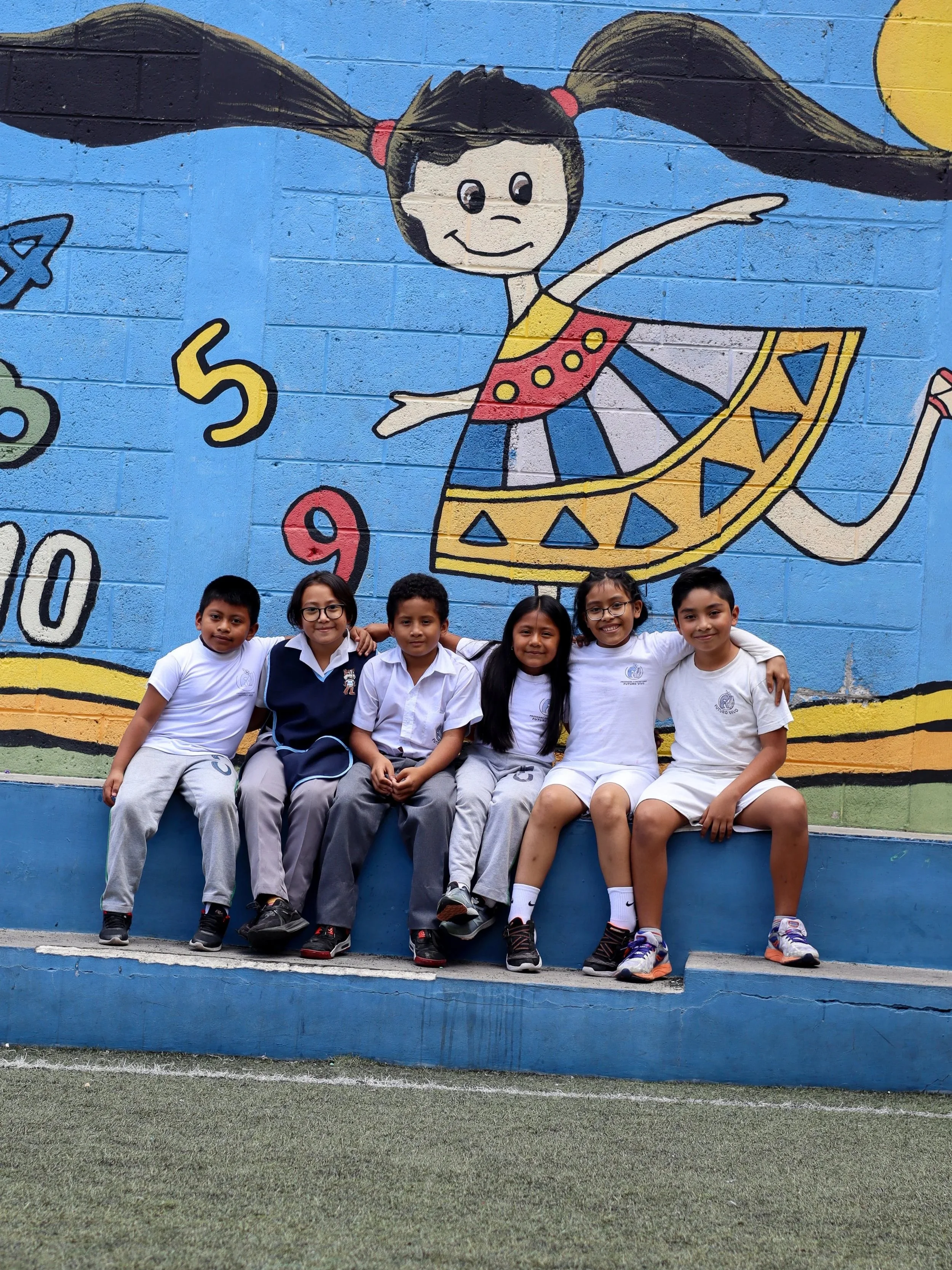 Grupo de seis niños sentados en un banco frente a un mural de una niña con maquillaje de payaso, vestida con una falda colorida, en un espacio escolar.